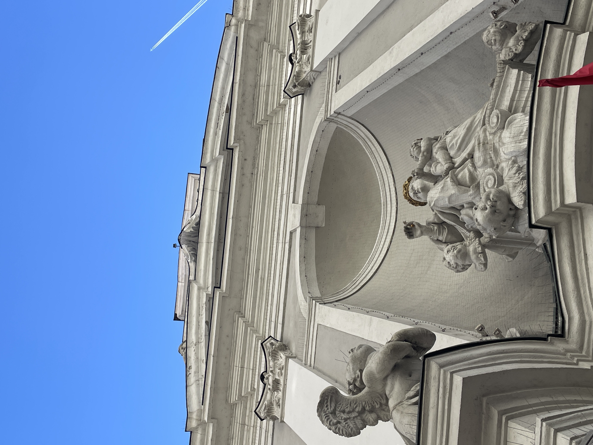 The image features a low-angle view of an ornate, light-colored building facade against a clear blue sky. A jet contrail cuts across the sky in the upper left corner. The building's facade is highly detailed, with vertical columns, moldings, and sculptural elements. A niche contains a sculptural group of figures including Mary and the Christ child, surrounded by cherubs. A fine mesh is also visible, apparently to protect the statues. The building is likely the Matthias Church located in the Castle District of Budapest, Hungary. The picture is taken from below at an angle and is oriented vertically.