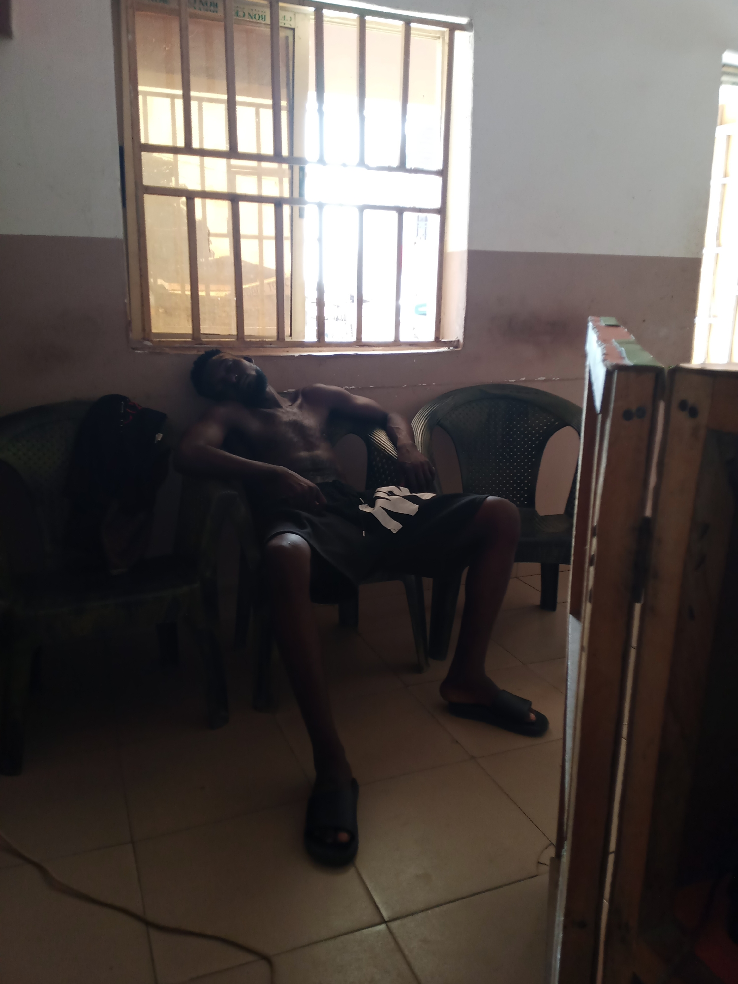 In an indoor scene likely from Jalingo, Nigeria, a man is asleep on a green plastic chair. He is shirtless, wearing dark shorts, and black sandals. His body is reclined, with his head resting against the back of the chair. Beside him, another green plastic chair is partially visible, and on it rests a dark object, possibly a bag. The environment features a large window with metal bars and tinted glass, allowing sunlight to stream in and create shadows on the tiled floor. The walls are painted in two tones, a lighter color at the top and a darker brown below. To the right, a wooden structure, possibly a shelf or a partition, stands. The lighting is dim, with strong contrast between the sunlit areas and the shadows.