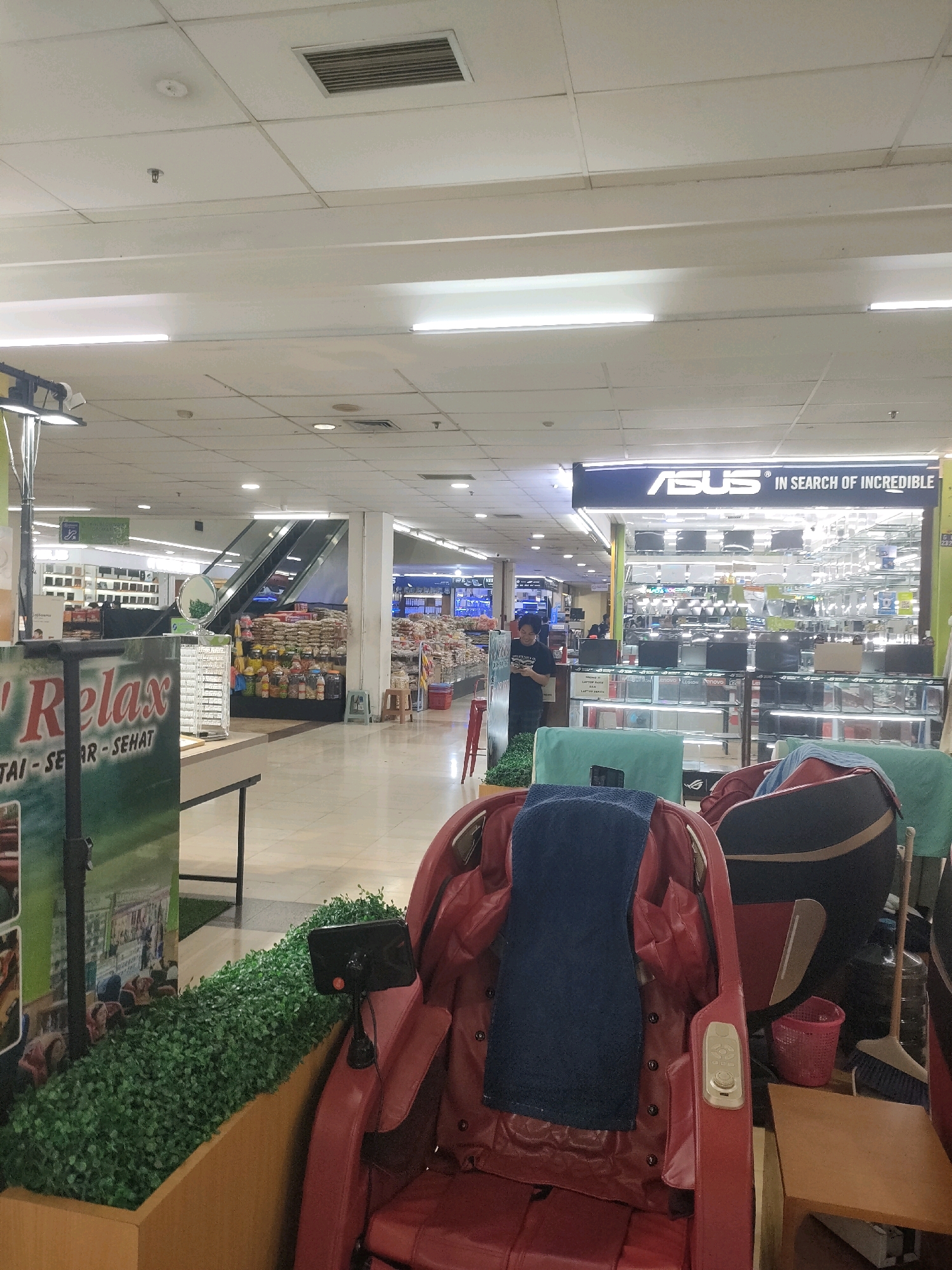 The image depicts the interior of a mall in Batam, Indonesia. In the foreground, there are two massage chairs, one red and one black and blue, positioned near a planter filled with artificial greenery. The red chair has a dark blue towel draped over its headrest and a control panel on its side. Behind the chairs, a sign that reads "Relax" is visible. To the right of the chairs, a small wooden table, a pink basket, and a broom are present.
Further back, a wide, brightly lit mall corridor stretches out. Escalators are visible on the left, leading to an upper level. Various shops line the corridor, including an ASUS store with a prominent banner that says "ASUS IN SEARCH OF INCREDIBLE." The store displays numerous electronic devices, likely laptops and monitors. A person in a black shirt stands near the ASUS store, looking at their phone. Shelves stocked with goods are visible in other shops, suggesting a retail environment. The ceiling features fluorescent lighting and air vents. The overall environment is a typical indoor shopping mall setting.