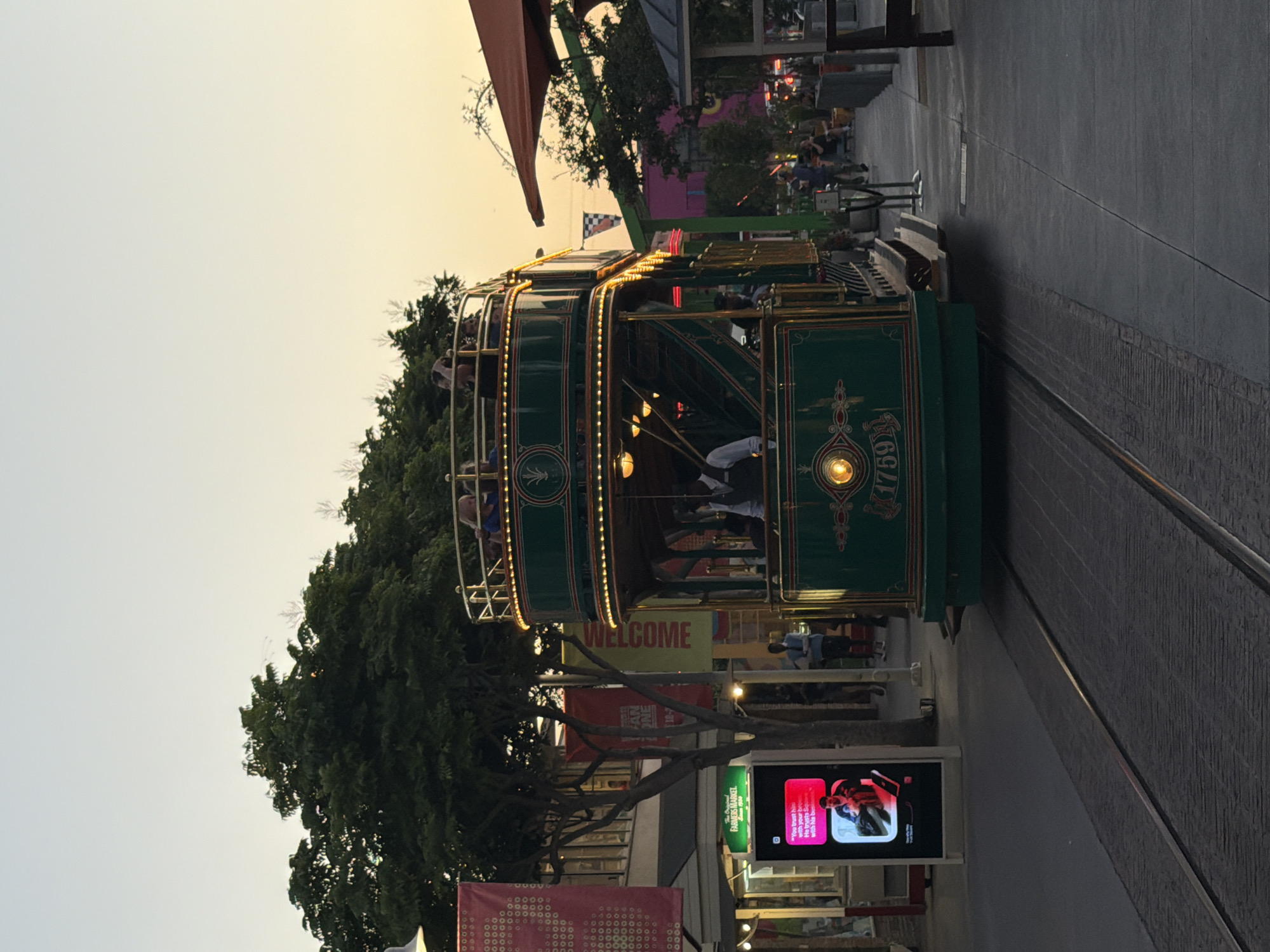 A double-decker green and gold trolley car is on tracks in what appears to be a pedestrian area of The Original Farmers Market in Los Angeles. The trolley has people visible on both levels. A digital advertisement screen displaying a man is to the left of the trolley. Several trees and buildings are behind the trolley, with a clear sky above, suggesting it is either early morning or late afternoon.