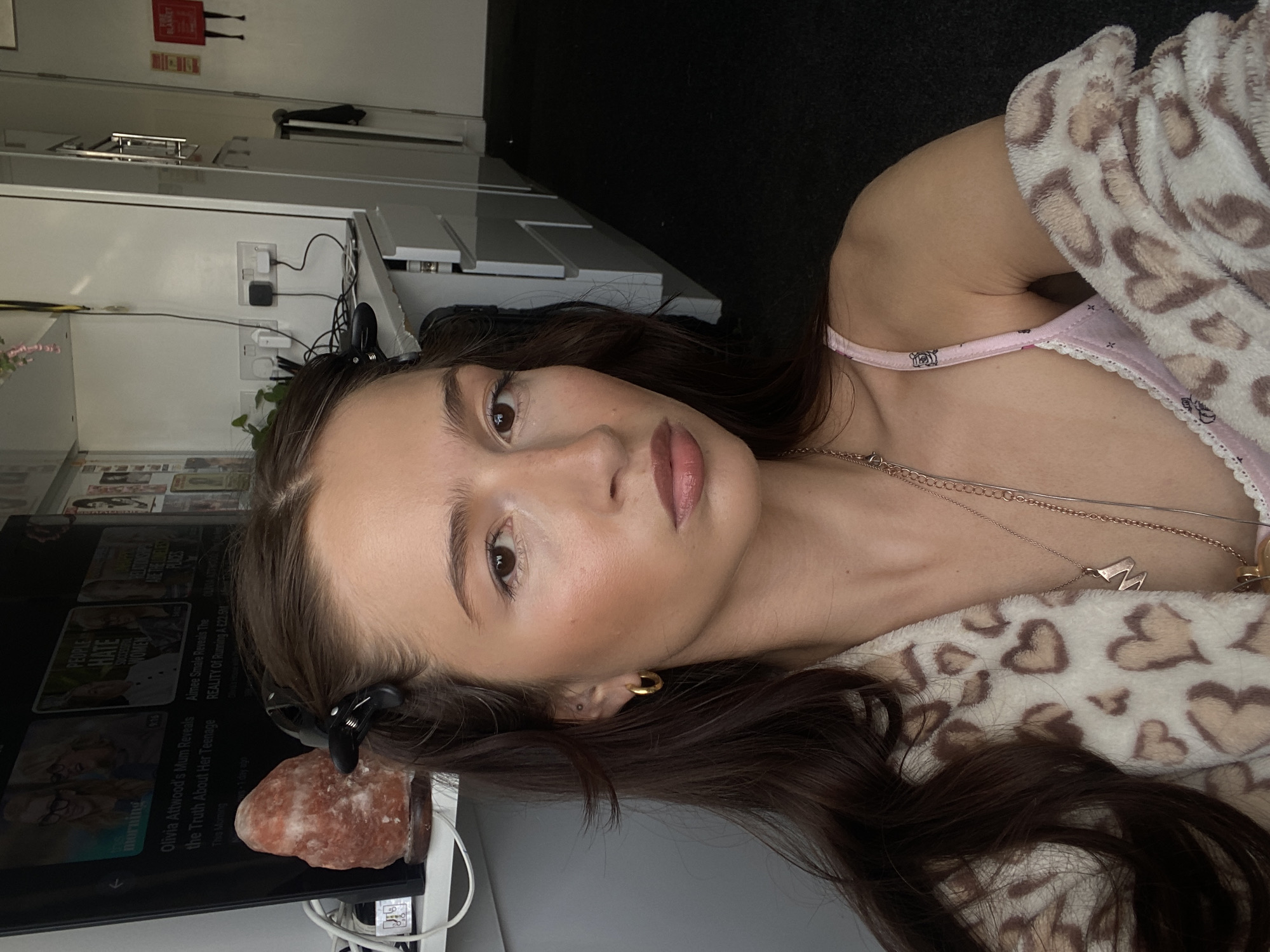 A young woman with dark brown hair and full lips is looking at the camera. She is wearing a light pink camisole with a lace trim and a fluffy robe with a leopard print pattern. She has on gold hoop earrings and a necklace with a pendant. Her hair is pulled back with black clips on top. In the background, a television screen displays a morning show. A pink Himalayan salt lamp is on a shelf next to the television. The woman is in a room with white walls and dark carpet. Electrical outlets and cords are visible on the wall behind her, along with some shelving and what appears to be a cabinet. The lighting suggests it is daytime. The scene is likely a domestic setting within London, United Kingdom.