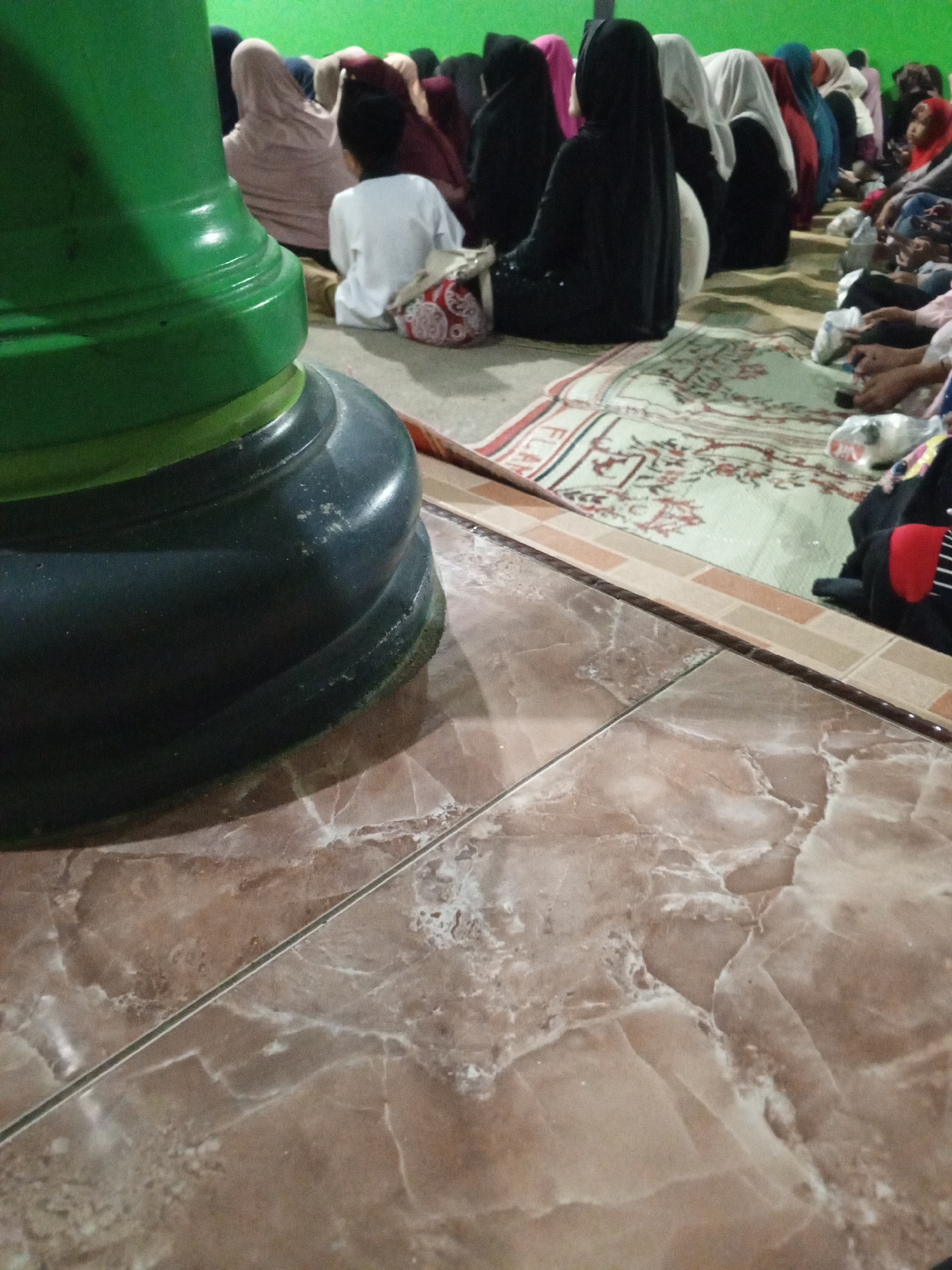 The image depicts a group of people, predominantly women and at least one child, sitting on the floor, facing away from the camera. They are dressed in traditional Islamic attire, with most of the women wearing hijabs and abayas in various colors, including black, pink, white, and red. A young child in a white shirt and dark pants is also visible, sitting on a patterned cushion. The setting appears to be an indoor space with a green wall in the background and a floor covered with a patterned rug. A large, dark green, cylindrical object is prominently positioned in the foreground, partially obscuring the scene. The floor in the foreground is tiled with a brown and beige marble-like pattern. The image was taken in Lemahbang, Indonesia. The people appear to be engaged in a religious activity or gathering.