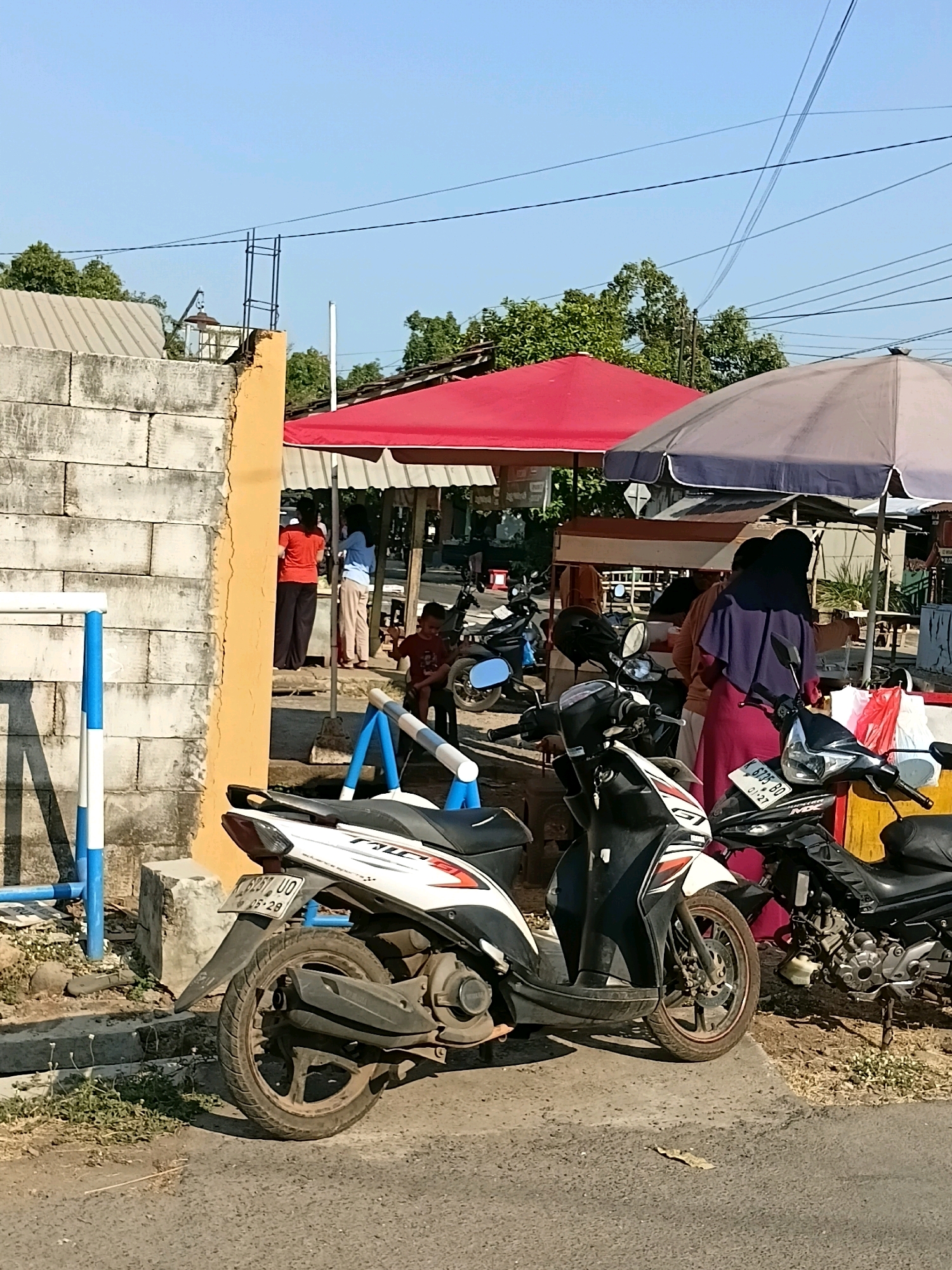 This is a daytime, eye-level, medium shot of a street scene in Cikal, Indonesia. The image captures a row of parked motorcycles, some with people standing or sitting around them. Two food stalls are visible, one with a red canopy and another with a grey umbrella. The stalls appear to be set up along a street or sidewalk, with a rough-looking wall on the left side of the frame. A young boy is seated on a chair near the stalls, and several adults are engaged in conversations or transactions. The overall atmosphere suggests a typical bustling street market or roadside eatery.