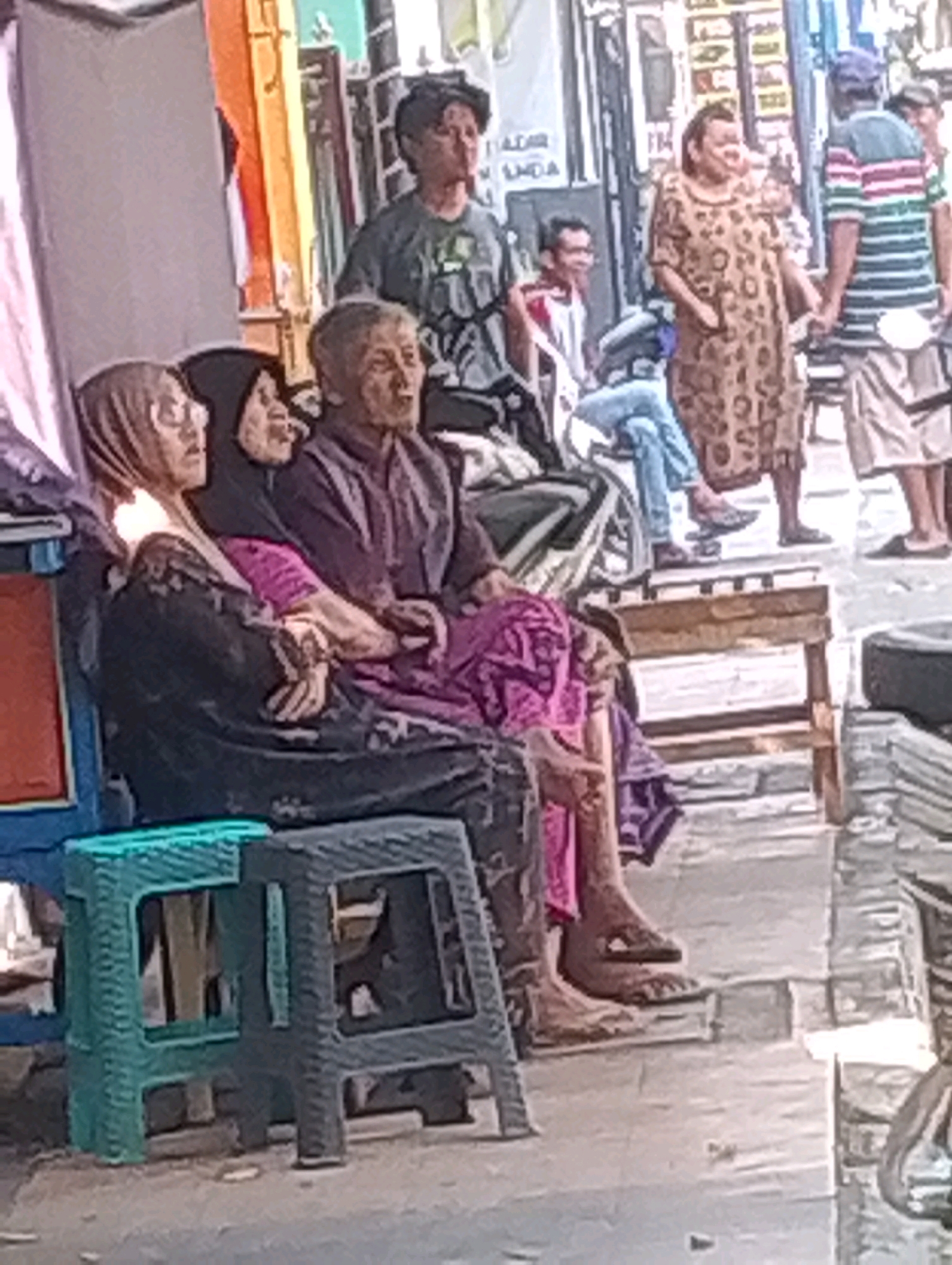 In Probolinggo, Indonesia, a group of individuals are gathered outdoors, with some seated on plastic stools. In the foreground, two women and an elderly man are seated on dark and teal plastic stools. The woman on the left wears a hijab and glasses, and is dressed in a dark outfit. Next to her, another woman wears a black hijab and a purple garment. The elderly man beside them is dressed in a purple shirt. Further back, another man is visible, as well as a woman in a patterned dress and a man in a striped shirt and shorts. The setting appears to be a street or market area, with buildings and stalls in the background, suggesting a bustling urban environment.
