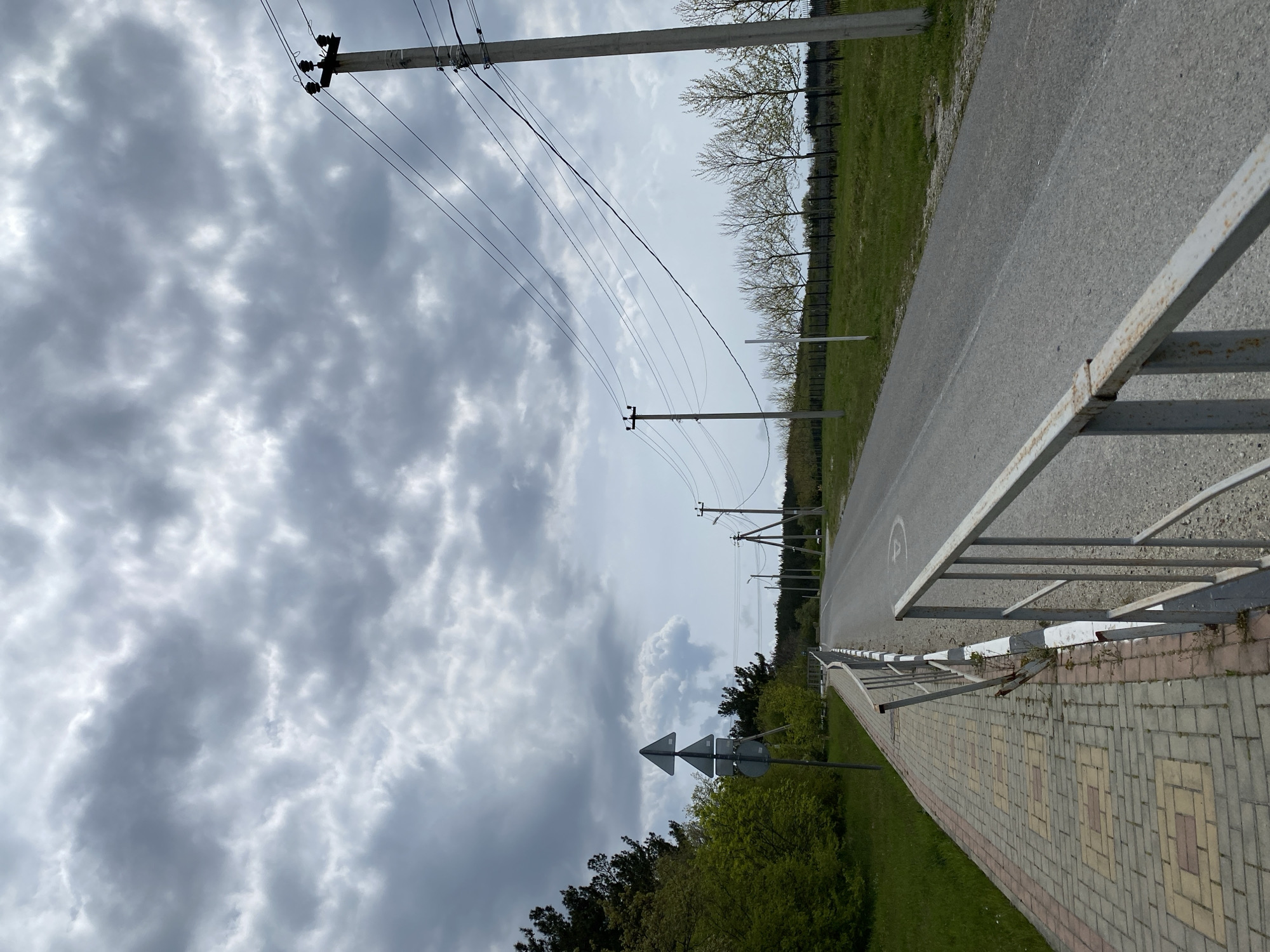The image depicts a rural or semi-rural roadside scene. A paved road stretches into the distance, bordered by a metal guardrail and a brick-paved sidewalk on one side. Power lines are visible overhead, supported by utility poles. The sky is overcast with heavy clouds. Vegetation, including trees and bushes, lines the sides of the road. A road sign with directional arrows is present. The precise location within Gelendzhik, Russia, cannot be determined from the image alone.