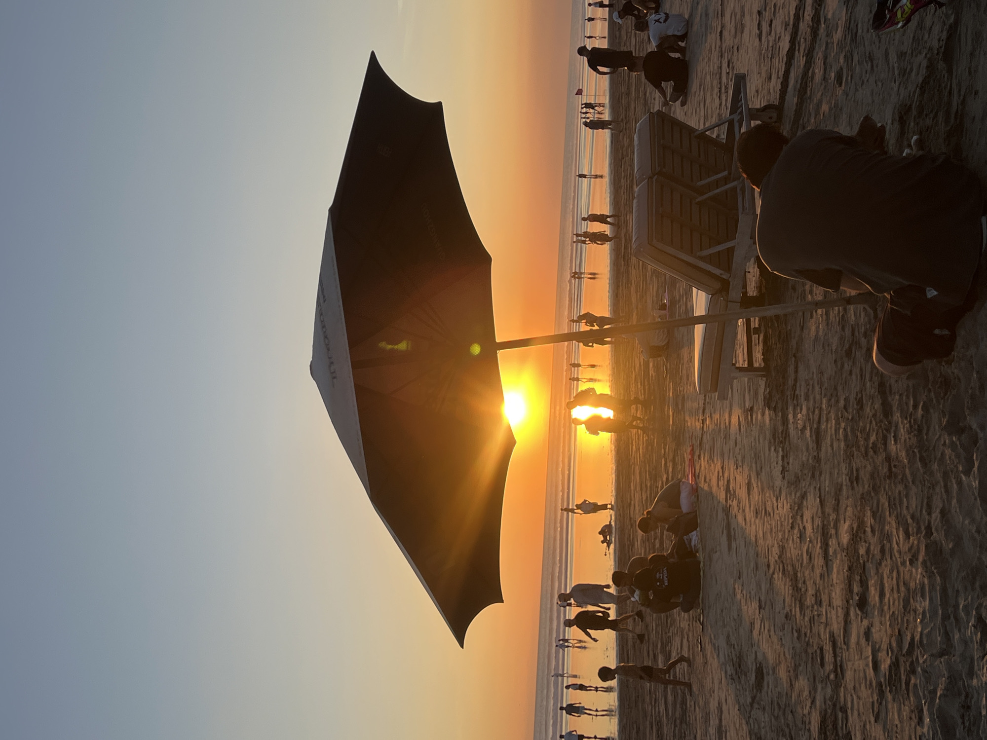 This photograph captures a vibrant sunset scene on a crowded beach in Kuta, Indonesia. The sun, a brilliant orange-yellow orb, is positioned low on the horizon, creating a powerful lens flare and bathing the sky in warm, radiant hues that transition from a soft blue at the top to a deep golden orange.
In the left foreground, a large, dark beach umbrella stands out, with the word "SUNZSHINE" faintly visible on its surface. To the right of the umbrella, a person in a dark t-shirt and shorts is seen crouching on the sandy beach, with their back to the camera, observing the sunset.
The beach is populated by numerous individuals, mostly silhouetted against the bright sky and the shimmering water. People are scattered across the sand, some standing, some sitting on beach chairs or directly on the ground. Their presence suggests a lively, communal gathering to witness the evening spectacle. The wet sand near the water reflects the vibrant light and some of the figures. Faint text, possibly "SOPEDA," is visible on the back of one person's dark shirt.
The clear sky and calm ocean indicate pleasant weather. The overall atmosphere is one of peaceful enjoyment and appreciation of the natural beauty of a tropical sunset.