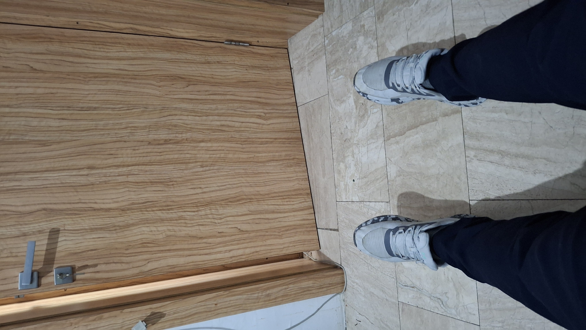 The image shows a person's legs and feet wearing grey and white athletic shoes and dark blue pants, standing on a tiled floor in front of a wooden door. The door has a silver handle and a visible lock. The floor tiles are light beige with subtle brown marbling. The wooden door has a distinctive vertical grain pattern. The scene appears to be indoors, likely in a residential or commercial building. The image does not provide any specific details about the city of Nesebar, Bulgaria, or any context that would place it there.