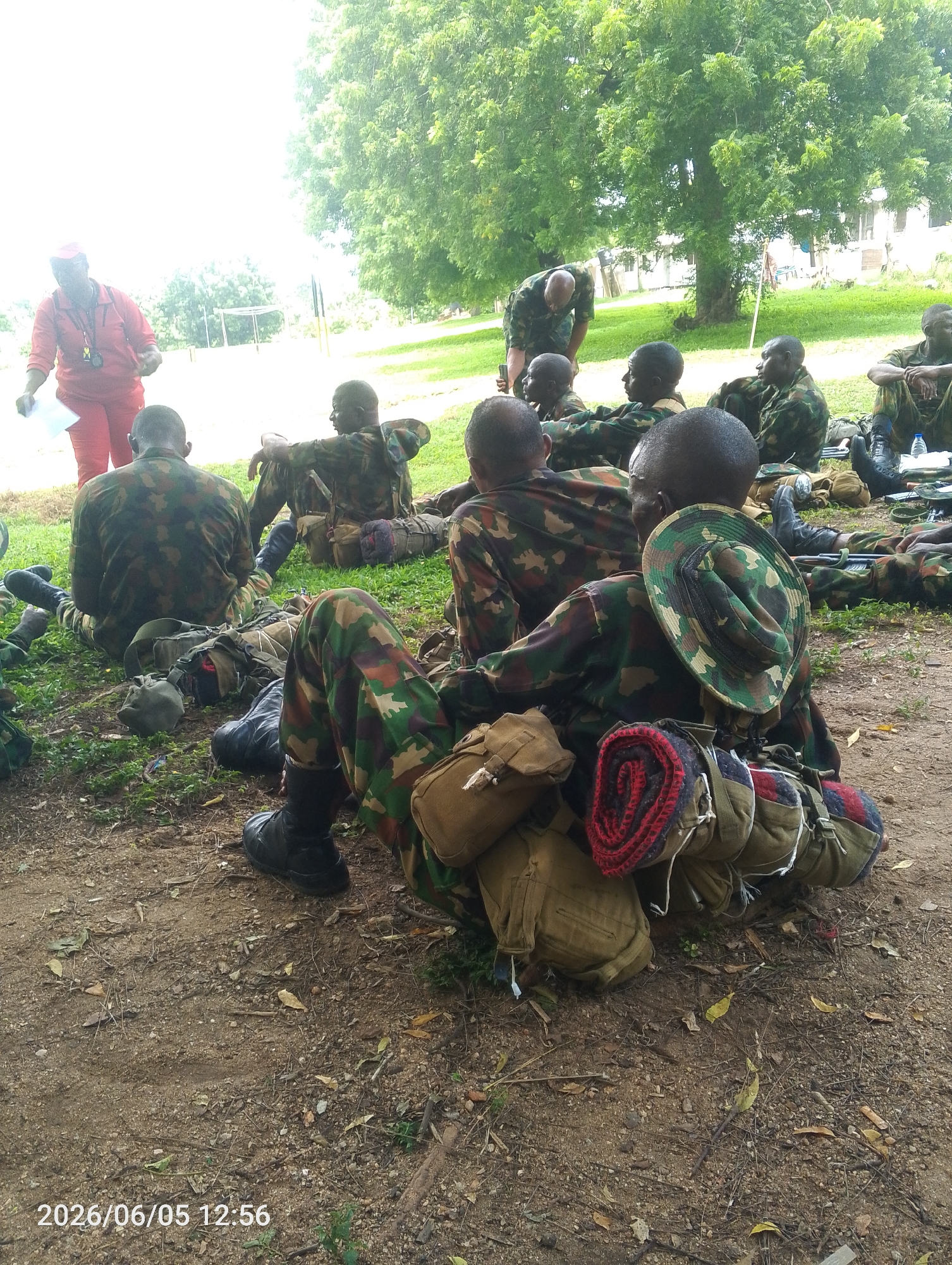 This outdoor image, taken in Adankolo, Nigeria, depicts approximately a dozen individuals, primarily men in camouflage military uniforms, gathered under large green trees. Most are seated on the ground, which is a mix of dusty earth, small rocks, and sparse green vegetation. Their military equipment, including backpacks, canvas bags, and a rolled-up red and black blanket, lies beside or on them.
A woman in a red tracksuit and cap stands to the left, holding papers and wearing a whistle around her neck, suggesting an instructor or official role. Another individual in camouflage is seen bending over a seated group further back, possibly also in an instructional or supervisory capacity. The scene appears to be a training session, briefing, or a rest period during a military exercise.
Notable details include black military boots worn by the soldiers and a camouflage bucket hat resting on one soldier's equipment in the foreground. In the background, beyond the trees, distant buildings are faintly visible. The lighting indicates bright conditions, consistent with the midday timestamp "2026/06/05 12:56" prominently displayed in the bottom-left corner of the image. The overall mood appears focused and calm, with individuals either listening attentively or resting.