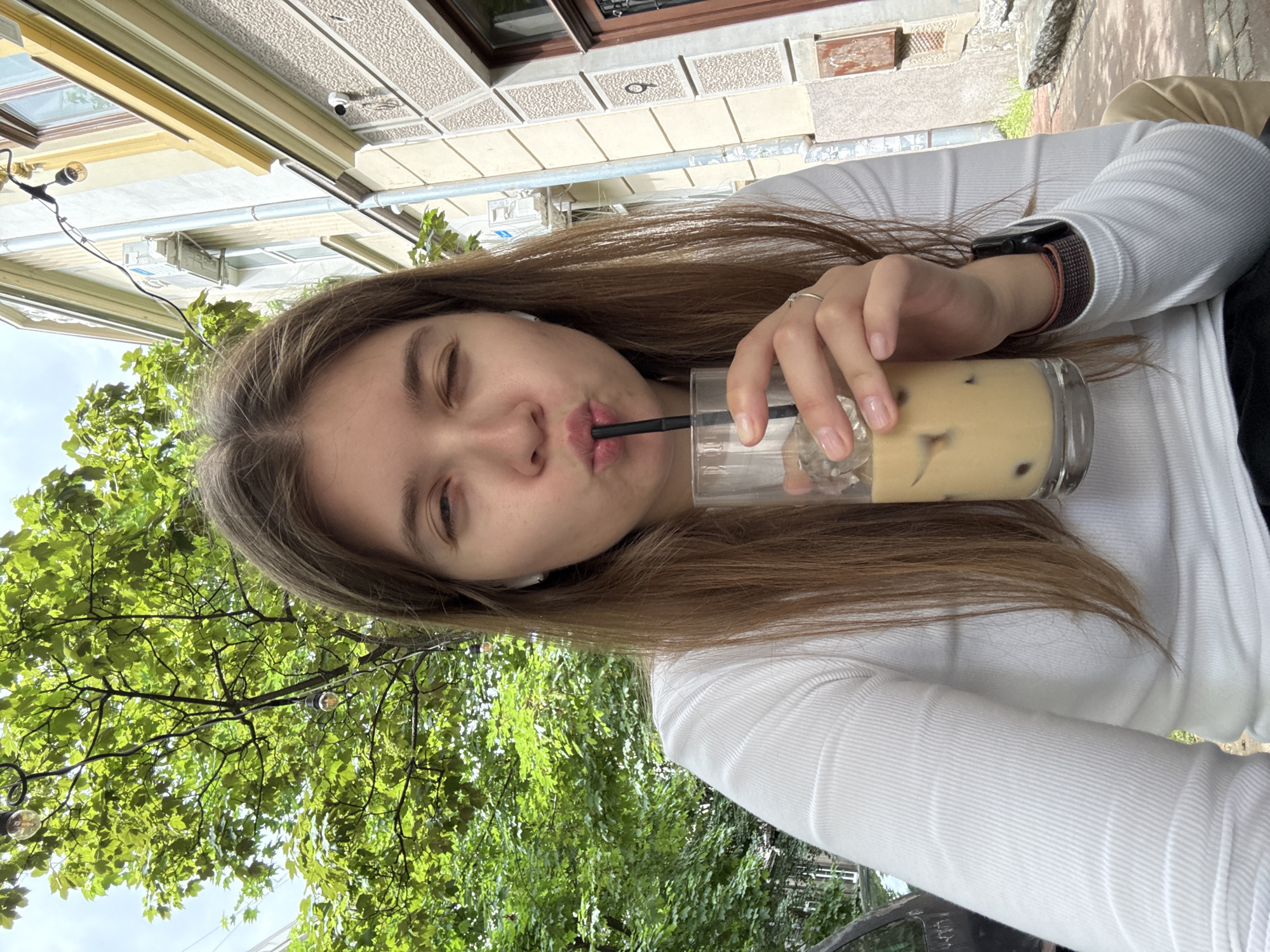 A young woman is drinking from a glass filled with a tan-colored beverage and ice through a black straw. She is wearing a white long-sleeved shirt and has a watch on her left wrist. The woman is outdoors, likely at a cafe or restaurant in Belgrade, Serbia, judging by the architecture in the background. A building with beige trim and a security camera mounted on the facade is visible behind her. Lush green trees and decorative string lights are present, suggesting a pleasant outdoor setting. The image appears to be a selfie taken from a slightly upward angle.