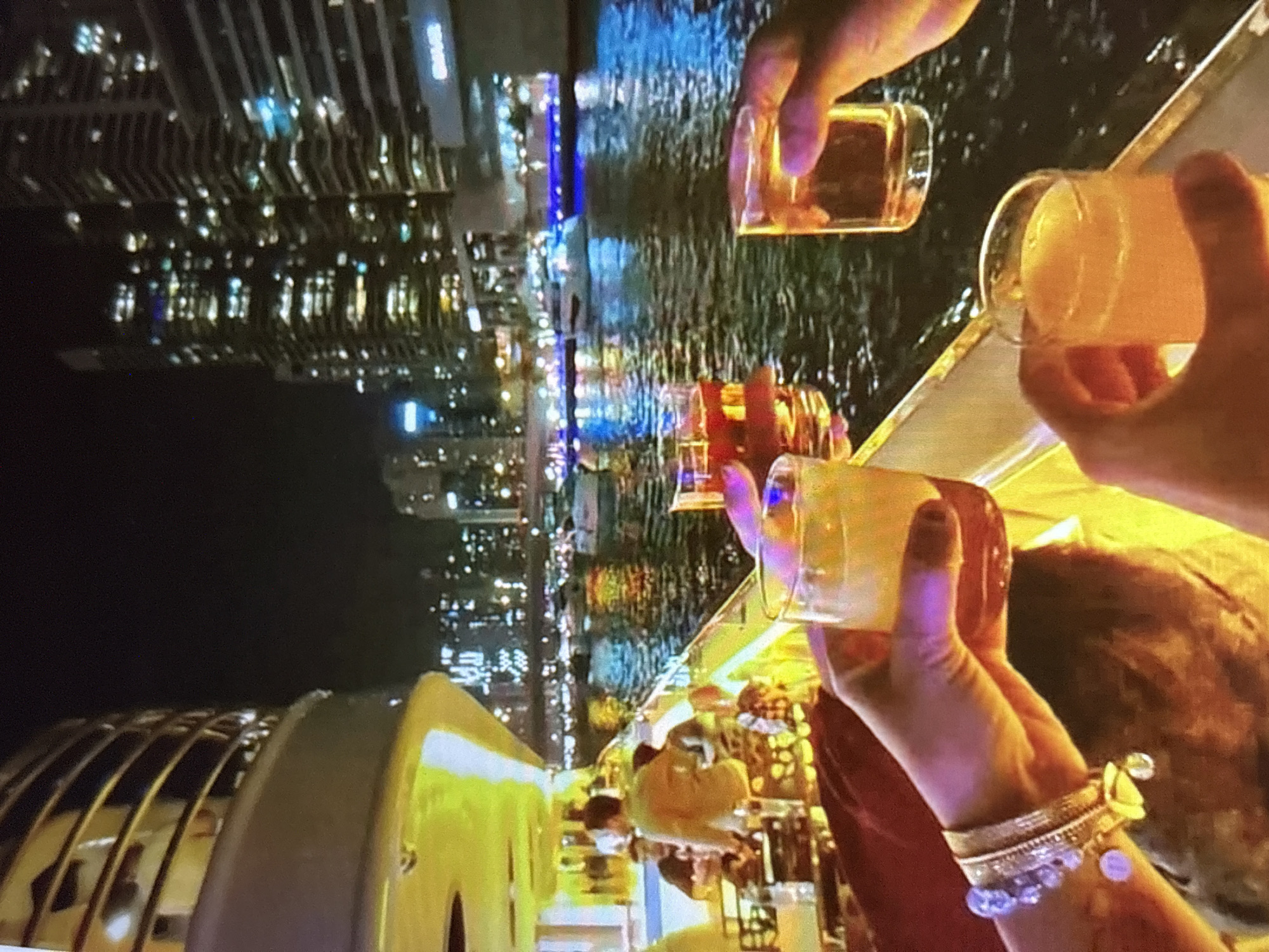 In this media file, a group of people are enjoying drinks at night, likely on a boat or a waterfront venue, given the reflections of city lights on the water. The scene is illuminated by artificial lights from the city skyline and some internal lighting.
The primary subjects are hands holding clear glasses filled with amber and yellowish beverages. One hand wears an ornate bracelet with charms, suggesting a celebratory or social occasion. In the background, out of focus, are glimpses of other people and what appears to be a boat's railing or seating area. The reflections in the water suggest a metropolitan setting with tall buildings.
The activity is a toast or a casual gathering where drinks are being shared. The time of day is clearly night, with the city lights creating a vibrant backdrop. The mood appears relaxed and social. The location context is Pori, Finland, which might imply a summer evening given Pori's known festivals and long daylight hours during summer. However, the image itself predominantly shows nighttime. There is no visible text in the media file.