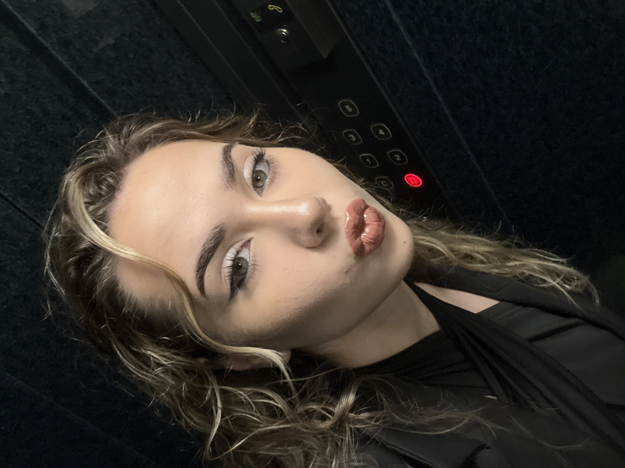 A woman is taking a selfie in an elevator. The elevator control panel is visible behind her, with buttons numbered 1 through 5, and the button for the second floor is illuminated. The woman has light brown hair with blonde highlights, fair skin, and is wearing a black top or jacket. She is making a kissing face toward the camera. The background of the elevator appears to be dark. The location is unknown.