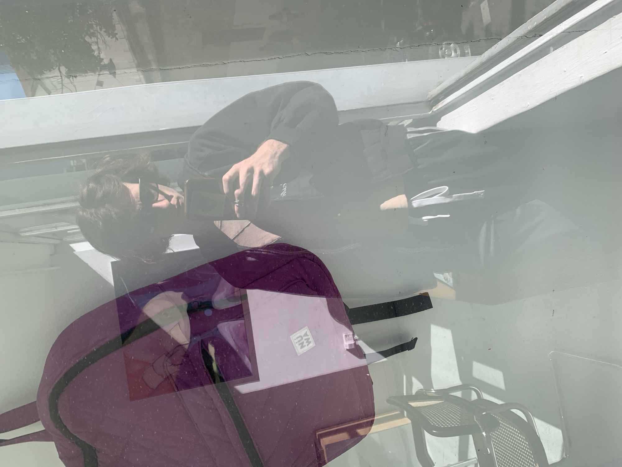 The image, presented rotated 90 degrees counter-clockwise from a typical upright perspective, captures a person through a reflective glass surface, likely a window. The individual is depicted horizontally, wearing a dark hoodie and looking down at a smartphone held in their right hand. A ring is visible on their right ring finger, and they have short, dark hair.
In the foreground, a prominent purple, quilted backpack with black straps is visible, partially obscuring the person. A light-colored laptop, featuring a white sticker with the dark letters "NU MA" on its lid, rests on the backpack.
The glass surface creates a complex overlay of reflections, including tree branches, parts of a building exterior, and a bright blue sky, suggesting an outdoor environment beyond the glass. These reflections contribute to a somewhat hazy and layered visual effect. In the background, visible through the glass and behind the person, are two metal chairs with perforated seats and backs, hinting at a cafe or outdoor seating area. The scene is brightly lit by natural light, indicating it is daytime. The overall setting appears to be a casual public space in Buenos Aires, Argentina.