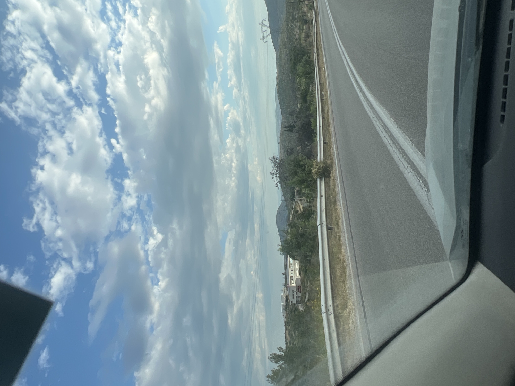 An outdoor, daytime, eye-level shot captures a view from inside a vehicle on a paved road. The road, marked with double white lines, curves slightly to the right. A metal guardrail runs along the left side of the road, separating it from a dry, grassy embankment. Beyond the embankment, the landscape unfolds with rolling hills covered in scattered vegetation and trees. In the middle ground, a modern, white building complex with several units and a red car parked in front is visible, situated amidst greenery. Further in the distance, a large, pylon-like structure with power lines is prominent against the sky, and undulating hills fade into the horizon under a partly cloudy sky. The sky itself is a vibrant blue, interspersed with scattered white and gray clouds. The lower portion of the image shows the interior of the vehicle, including the dashboard and windshield.