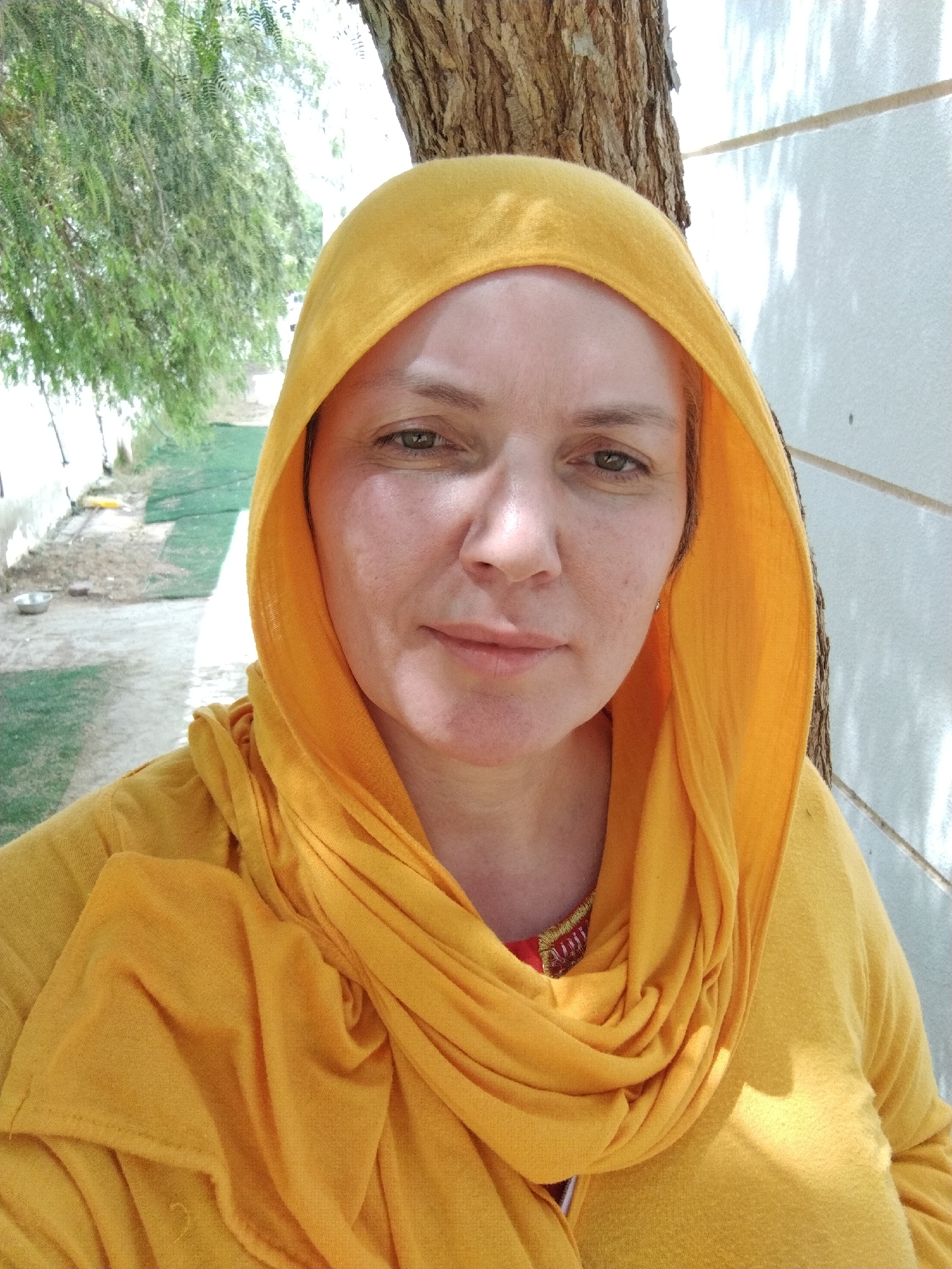 A woman wearing a yellow hooded shawl is standing outdoors. Behind her is a tree trunk and a white wall. To her left is a grassy area with a tree line in the distance. The image was taken in Al Mas'oudi, United Arab Emirates.