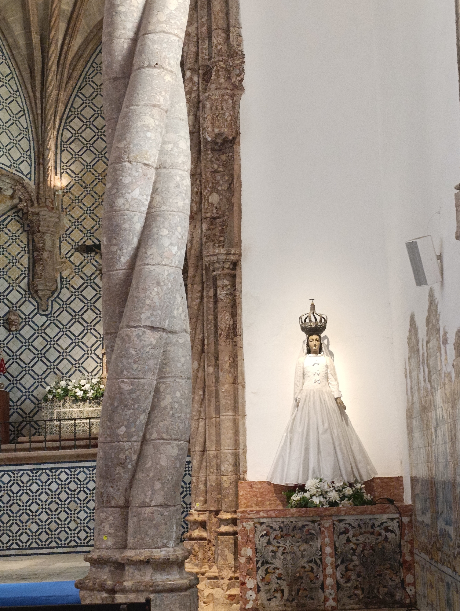 The image depicts the interior of a church in Setúbal, Portugal. In the foreground, a large, twisted stone column dominates the left side of the frame. Behind it, to the right, stands a statue of a woman wearing a long white dress and a crown. The statue is adorned with a wreath of white flowers at its base. The background features ornate tilework on the walls and a vaulted ceiling. The overall impression is one of religious and architectural grandeur.