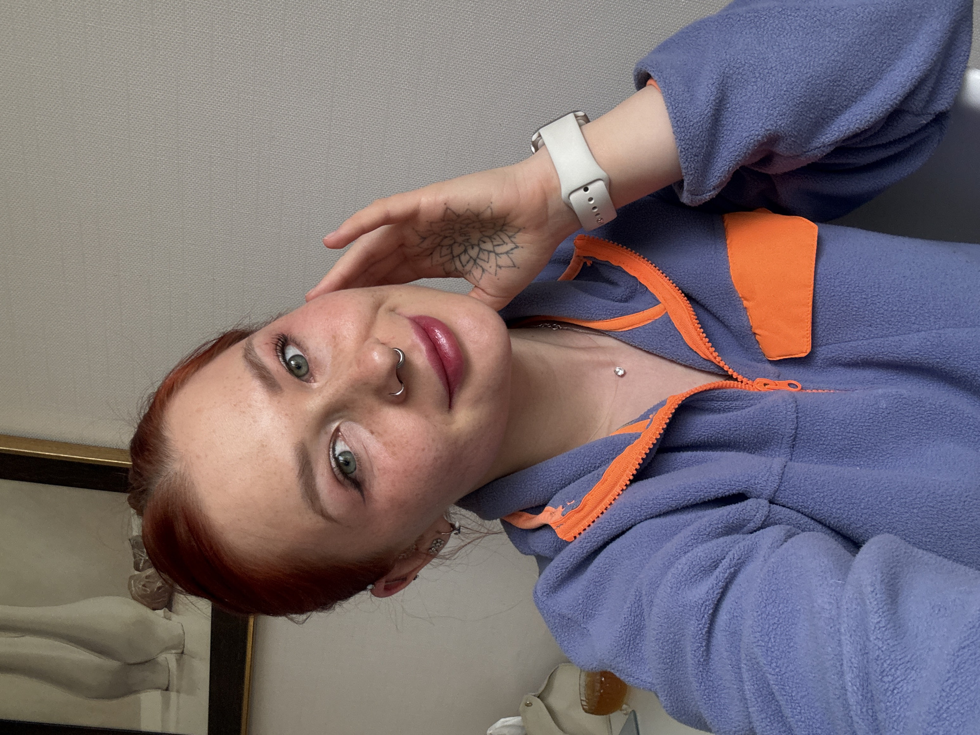 A young woman is captured in a close-up, rotated selfie, likely taken indoors in Rostov-on-Don, Russia. She has reddish hair, green eyes, and visible freckles across her face. Her lips are enhanced with a pinkish gloss, and her eyes are lined. She wears a silver septum piercing and what appears to be a dermal piercing on her chest. A black line-work tattoo, resembling a mandala, is visible on the back of her left hand, which is held near her face. On her left wrist, she wears a white smartwatch. She is dressed in a textured, purple fleece jacket with distinctive orange zipper, collar, and pocket details. Her expression is gentle and calm, with a slight smile as she looks towards the camera. The background features a light gray wall and, to the lower left, part of a framed artwork or sculpture depicting a classical-style white reclining figure. No text is visible.