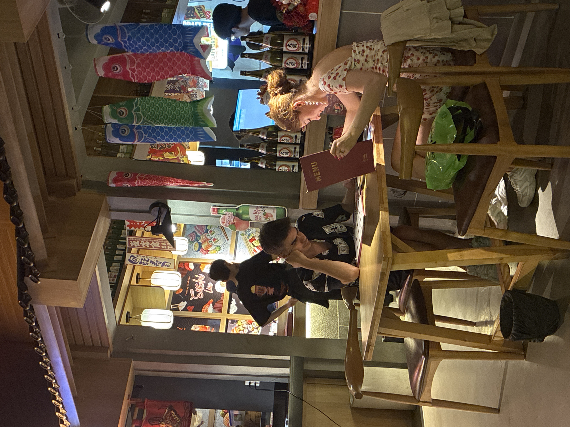 The image shows the interior of a restaurant, likely an eatery serving Japanese cuisine. There are several people present, including a woman in a cherry-printed dress, who is looking at a menu. A man with short hair, wearing a black shirt with dollar-bill designs, is leaning on a table nearby, seemingly waiting to order. Behind him, another person in a black shirt is standing. The environment is decorated with Japanese-themed elements such as koinobori (carp streamers) hanging from the ceiling and panels featuring food illustrations, specifically sushi. Shelves stocked with what appear to be sake bottles are also visible. Text on the panels includes phrases like "Sushi Line," "Tomapy Isahaya," and "Yoru Teishoku," suggesting the cuisine is Japanese. There's also the word "MENU" clearly visible on the menu card being held by the woman. The restaurant features wooden chairs and tables, contributing to a somewhat rustic, yet modern ambiance. The overall setting suggests a Japanese restaurant, possibly located in a city such as Đà Nẵng, Vietnam, given the blend of Japanese elements with a contemporary setting.