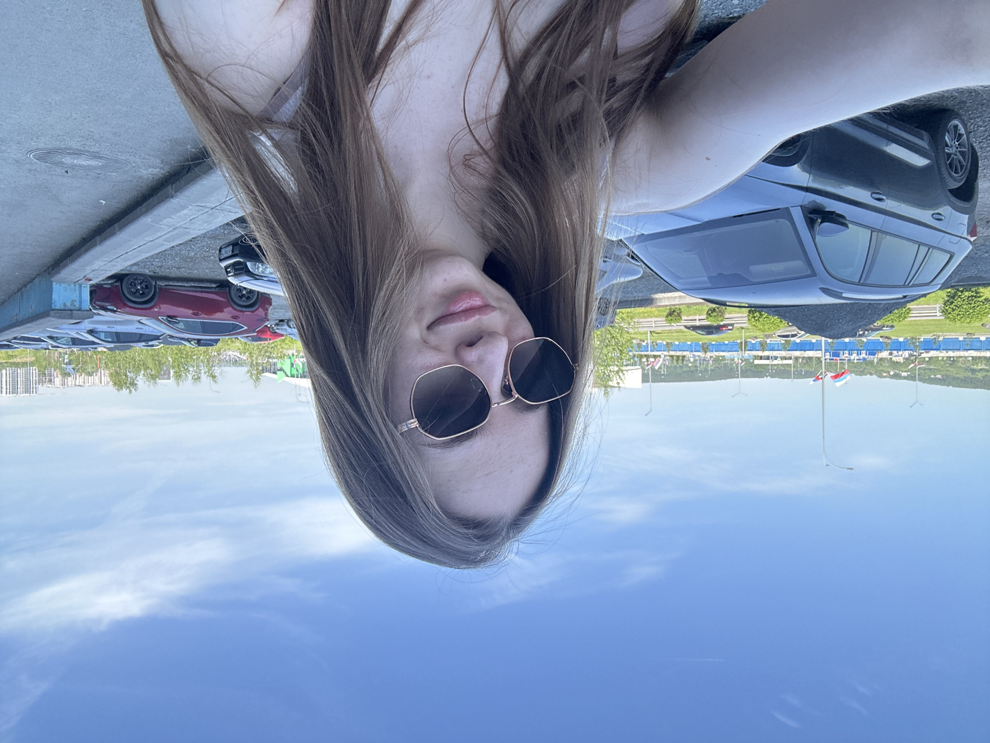 The image, correctly oriented, displays a close-up self-portrait of a young woman outdoors. She has long, straight, medium-brown hair that falls around her shoulders. Her eyes are obscured by dark, octagonal-shaped sunglasses with thin, gold-colored frames. Her lips are slightly parted, conveying a neutral expression.
The background reveals an outdoor setting, likely a parking area. Several cars are visible, including a dark grey SUV to her right and a red sedan to her left. Beyond the vehicles, there's a line of green trees and bushes, followed by a distant cityscape featuring numerous buildings. The sky above is clear and bright blue, with a few faint, wispy white clouds, indicating daytime. In the far distance, several flags on poles are visible; one clearly displays the white, blue, and red horizontal stripes of the Russian flag. Street light poles are also scattered across the distant landscape.
The scene suggests an urban or suburban environment, possibly a public parking lot or a viewpoint overlooking a city. The bright lighting and clear sky indicate pleasant weather conditions. There is no visible text in the image.