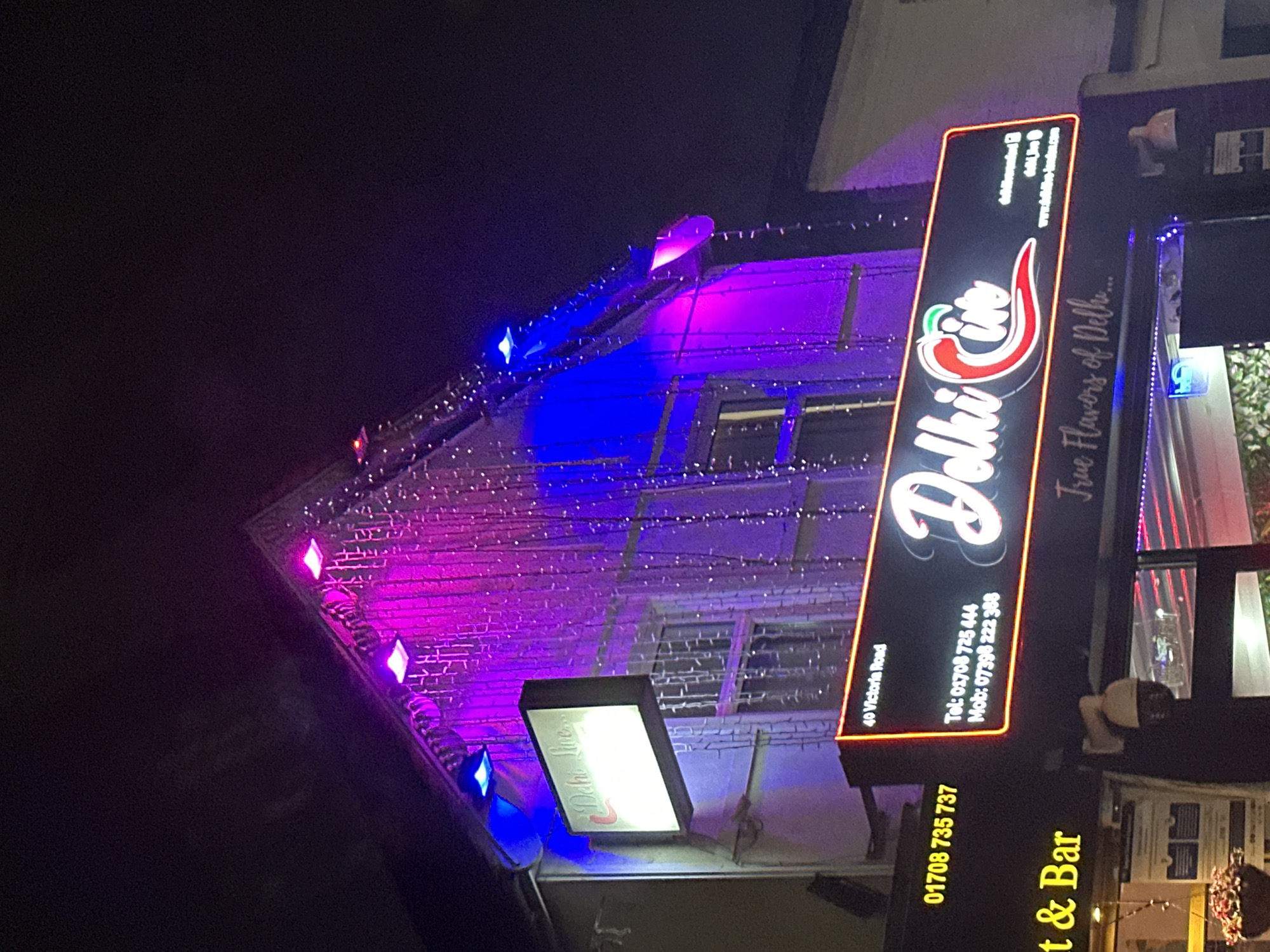 The image displays the illuminated exterior of a commercial building identified as "Delhi Cive" at 40 Victoria Road, Romford, London, United Kingdom, at night. The two-story building facade features a gabled roofline. The upper portion of the building is extensively decorated with cascading strands of white lights and is brightly illuminated by magenta and blue uplighting fixtures mounted along the roof and facade. A prominent rectangular black sign with a neon red border displays "Delhi Cive" in white script with a chili pepper graphic, alongside the partial text "True Flavors of Delh...". This sign also provides contact information: "Tel: 01708 725 444", "Mob: 07396 222 388", and website/social media details: "www.delhicive-london.com", "delhicivelondon", "delhicive_london". A smaller, illuminated rectangular sign above the main signage shows "Delhi Cive...". To the left, a partial vertical sign displays the number "01708 735 737" and the text "...t & Bar". A dome-shaped CCTV camera is mounted on the building below the main sign. A hanging plant container is visible in the lower-left foreground. No individuals or active interactions are depicted.