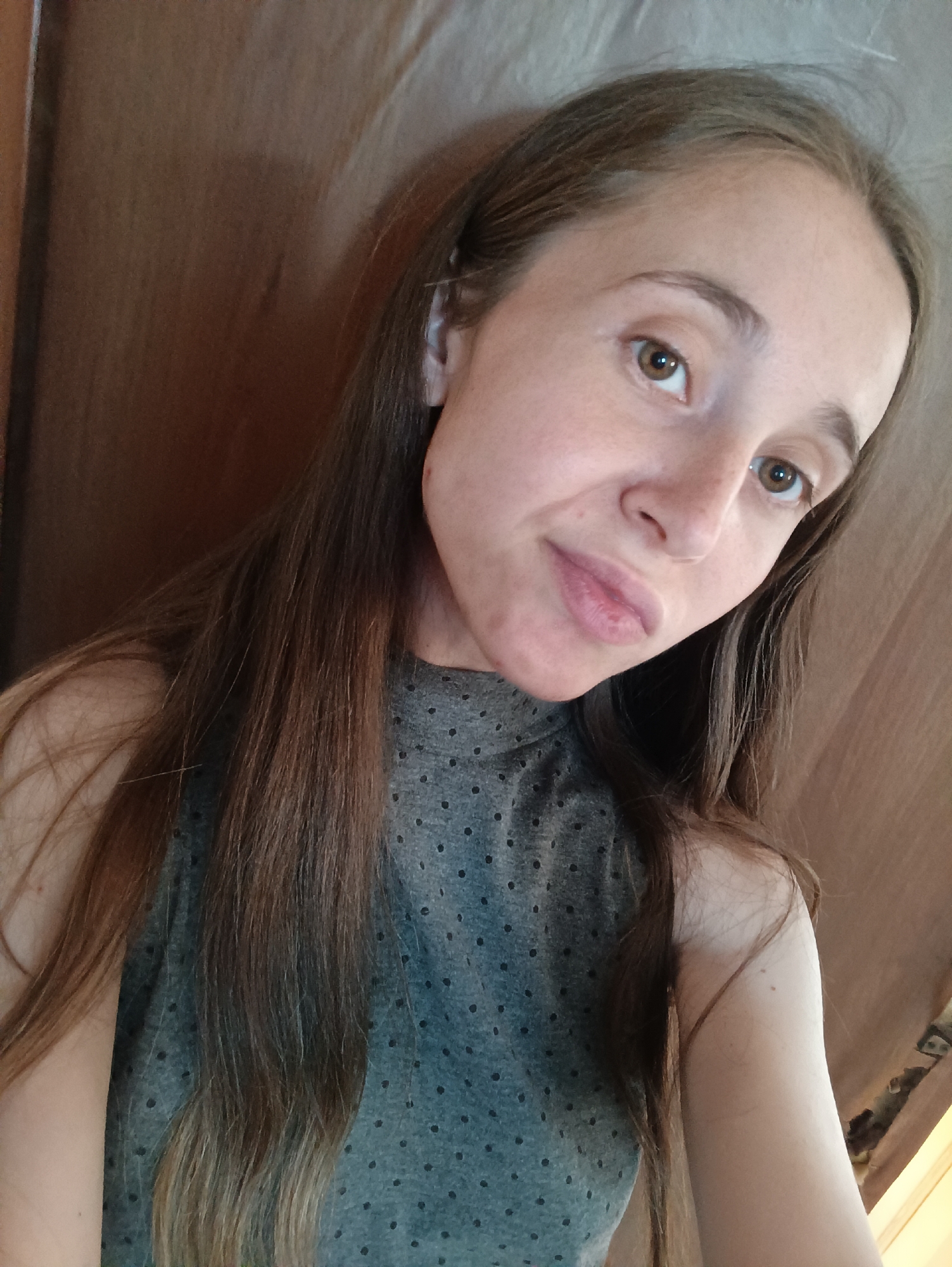 A medium shot shows a young woman with long, straight brown hair, and light brown eyes. She is wearing a gray, short-sleeved shirt with a high neckline adorned with small, dark dots. The woman is looking slightly to her right, with a subtle smile. Her skin is fair, and she has a few faint freckles across her nose and cheeks. The background appears to be a textured wall, possibly fabric, in a neutral beige color, with a wooden surface visible on the left side of the frame. The lighting is soft, casting a gentle illumination on her face. The image was taken outdoors in the city of Vypasne, Ukraine.