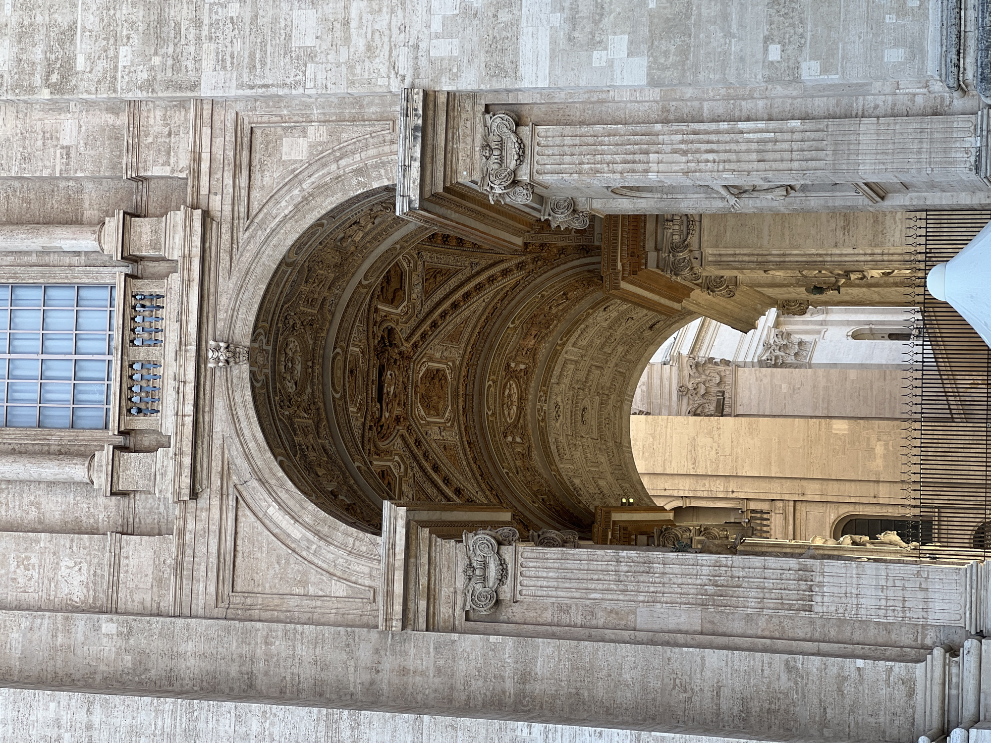 This image captures a close-up, upward-angled view of a monumental architectural structure, identified as part of St. Peter's Basilica in Vatican City, Italy. The dominant feature is a grand archway, constructed from light-colored, textured travertine stone, which recedes inwards, revealing an intricately carved and coffered ceiling. The ceiling details consist of ornate relief panels with patterns and rosettes, appearing in warm golden or light brown tones under direct sunlight.
The archway is flanked by engaged fluted columns or pilasters with elaborate composite capitals, featuring volutes and acanthus leaves. Above the arch, a large rectangular window with a light blue tint and a grid of panes is positioned. Below this window, a decorative balustrade with dark, stylized balusters is visible. A small, sculpted head or mask is integrated into the stonework directly above the apex of the main arch.
Strong, directional sunlight illuminates various surfaces, casting sharp shadows that accentuate the three-dimensional relief of the architectural elements and the deep texture of the stone blocks. Through the primary archway, another arch and further architectural details, including additional columns and sunlit walls, are visible in the background, suggesting a series of interconnected porticos or internal spaces. At the bottom right corner of the frame, a segment of a dark metal fence with vertical bars is present, along with a small portion of a white, conical structure, possibly a temporary canopy. No people are visible within the frame.