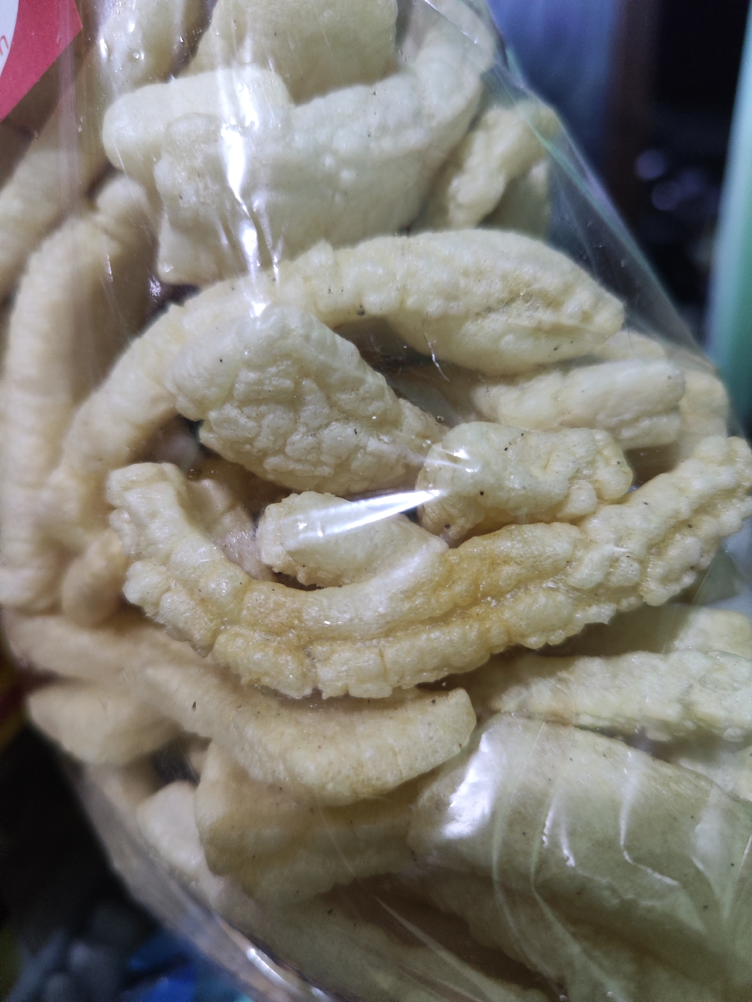 The image shows a close-up of a bag of fried snacks, likely fish crackers or similar, with a textured, puffed surface. The snacks are light yellow and appear to be piled loosely within a transparent plastic bag. Some of the snacks have a slightly golden-brown hue, indicating they have been fried. The lighting creates highlights and reflections on the plastic packaging. The background is out of focus, showing a dark, indistinct area with some faint blue and green hues. This image was taken in the city of Sroyo, Indonesia.