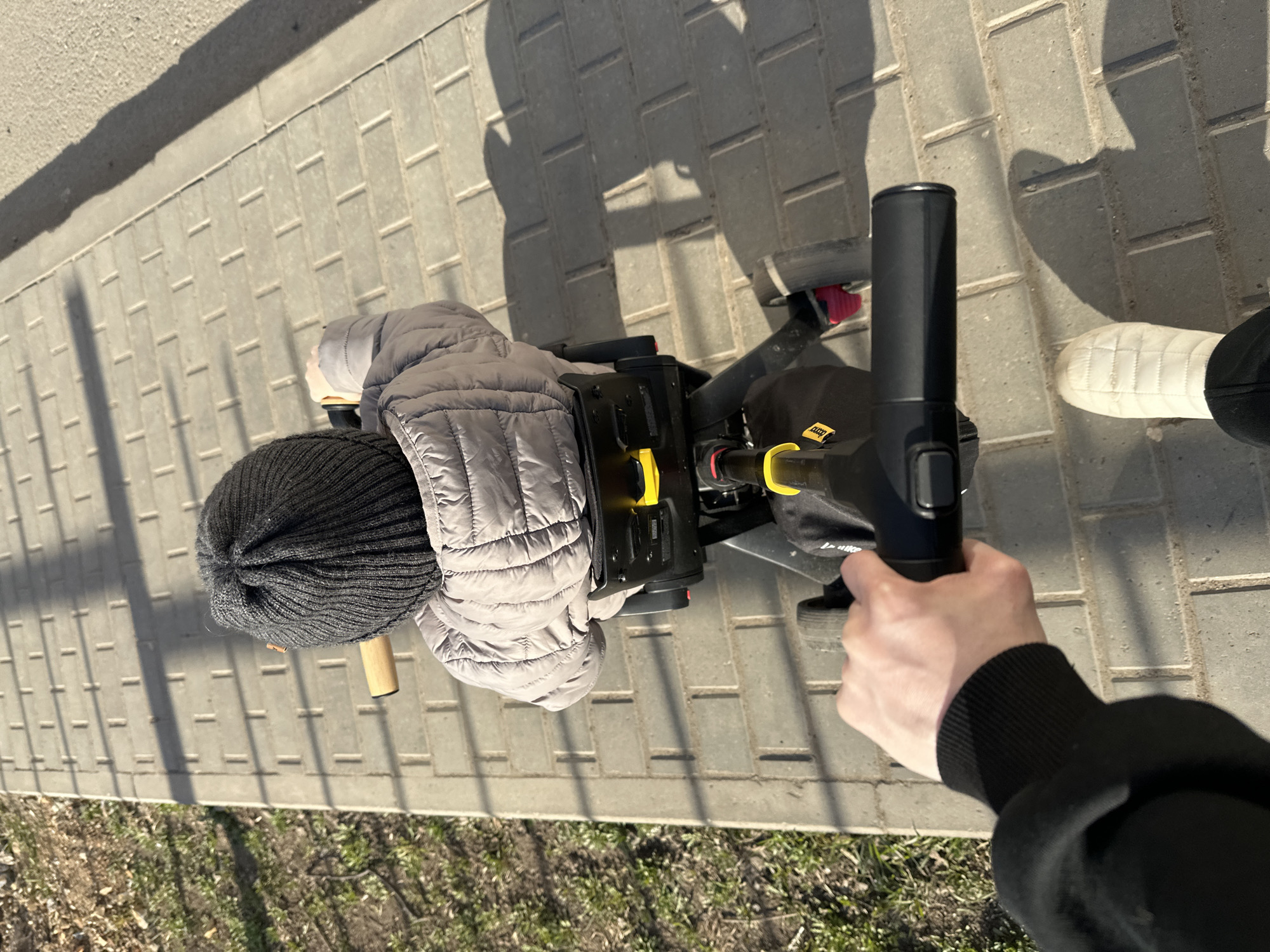 This aerial perspective image captures an adult pushing a child on a ride-on toy along a paved path in Votkinsk, Russia. The adult's right hand, wearing a black long-sleeved top with a ribbed cuff, firmly holds the black push handle of the toy. The adult's white quilted boot is visible on the right side of the frame. The child, viewed from behind, wears a warm grey puffer jacket and a black knitted beanie. The ride-on toy is predominantly black with bright yellow accents and includes a small black pouch bearing the white text "U-bike." Red components are also discernible on the toy's wheels or frame.
The outdoor setting features a meticulously patterned sidewalk constructed from rectangular grey pavers. Adjacent to the path, a strip of sparse, dry grass and soil is visible, suggesting early spring or late autumn. Strong, elongated shadows stretch across the paving, indicative of bright, direct sunlight, suggesting a clear day, likely in the morning or late afternoon. The scene portrays a casual outdoor activity.