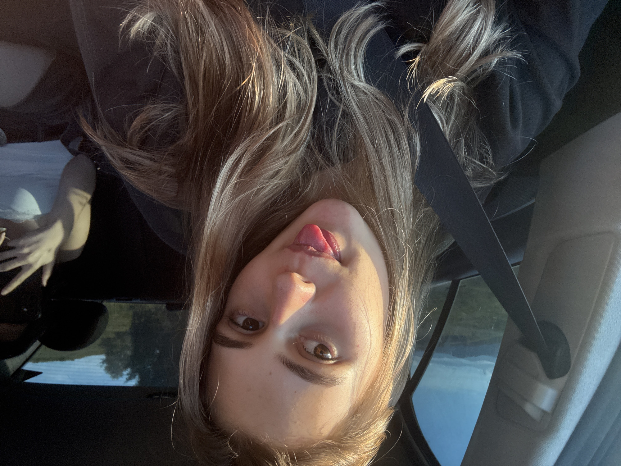 This is a close-up, portrait-style photograph of a young woman sitting in a car, taken from a high angle. The image is rotated 90 degrees counter-clockwise, so the woman's head is positioned on the right side of the frame, and her body extends towards the left. She has long, blonde hair that falls around her face and shoulders. Her eyes are open and looking towards the viewer, and her mouth is slightly open with her tongue sticking out playfully. The lighting suggests it is daytime, likely late afternoon, with warm sunlight illuminating her face from the left.
The woman is wearing a dark-colored top, possibly a hoodie, and a black seatbelt is visible across her body. In the background, through the car window, a landscape can be seen, with hints of trees and possibly water, suggesting an outdoor setting. The interior of the car is visible on the right side of the frame. The overall mood of the image is casual and lighthearted. The location is identified as Koprivshtitsa, Bulgaria, although there are no explicit text or visual cues within the image itself to confirm this.