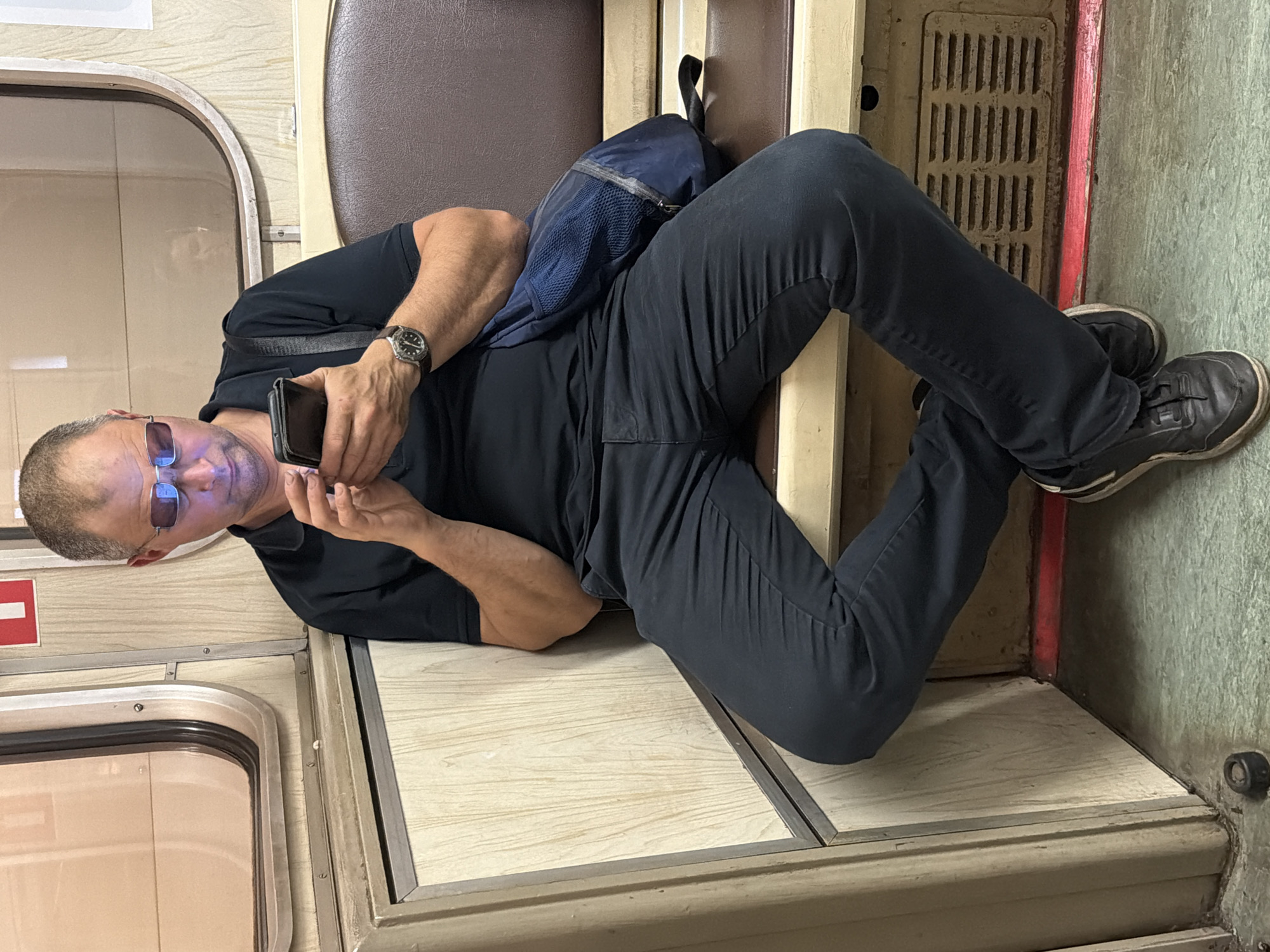 The media file shows a man lying on his side inside what appears to be a train carriage. The man is wearing a black t-shirt, dark pants, and dark sneakers. He has short, graying hair and is wearing sunglasses. He is holding a smartphone in his right hand and looking at it. A dark blue backpack is resting on his legs. The interior of the train carriage features light-colored wooden paneling and a dark brown seat. A metal grate is visible on the floor. The overall lighting suggests it is daytime. The presence of a train carriage interior and the specific visual style are consistent with Moscow, Russia. No visible text is present in the media file.