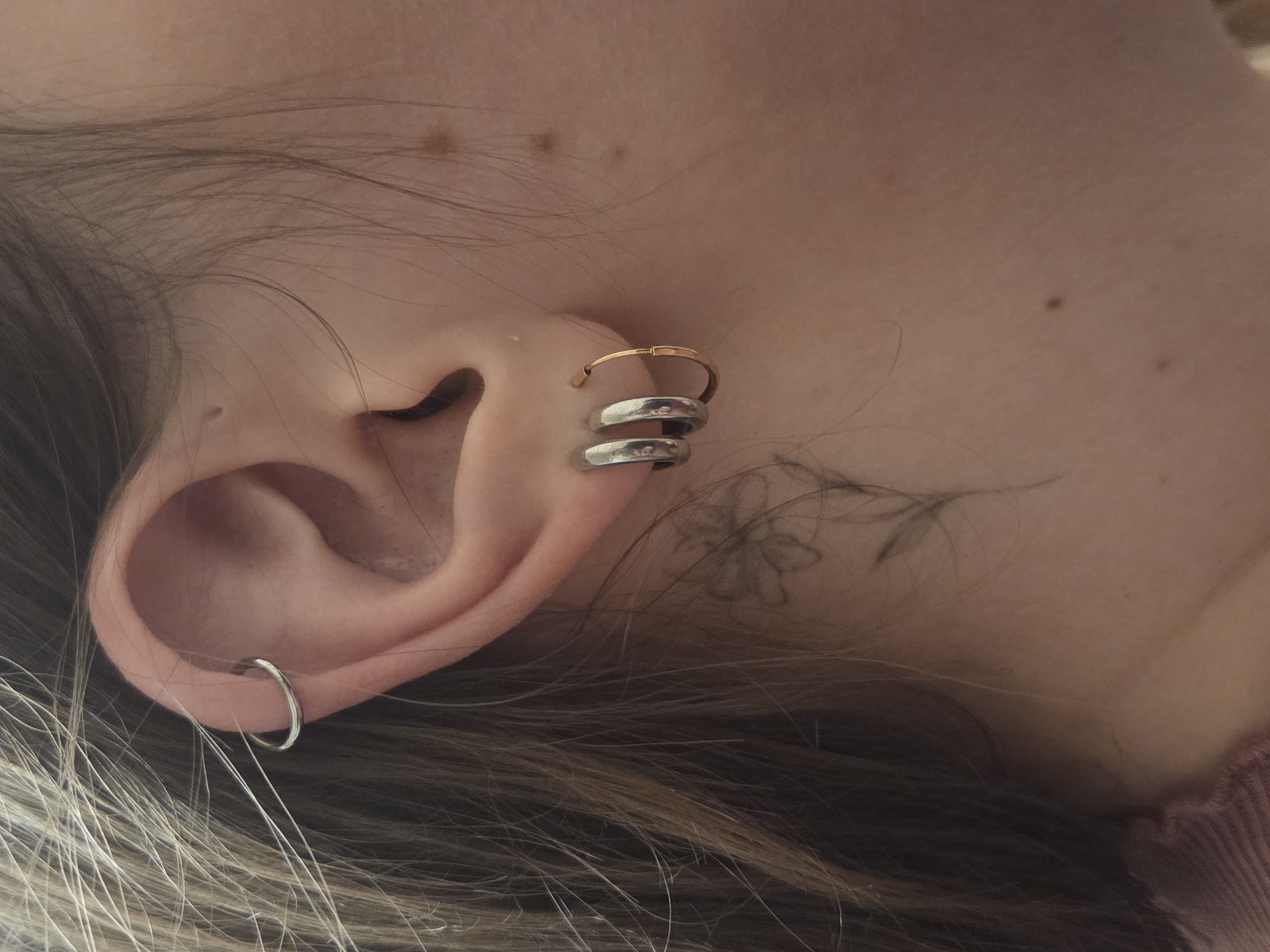 The image shows a close-up of a person's ear with several piercings. There is a small silver hoop in the lobe, and a gold hoop above it, and silver hoops around the helix. A faint flower tattoo is visible on the skin adjacent to the ear. The person has light skin and brown hair. The location is not discernible from the image.