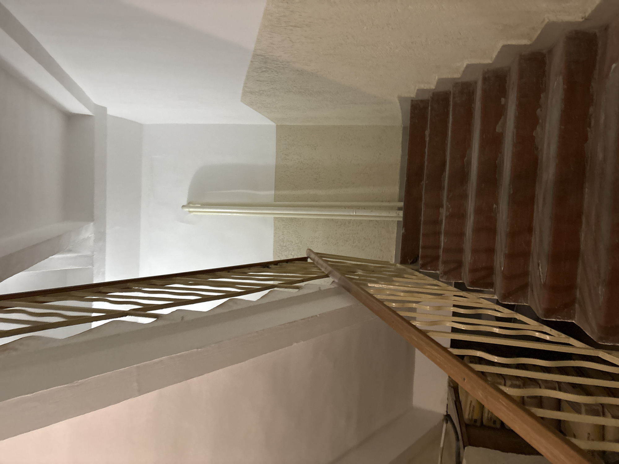 A view looking up a dimly lit, narrow apartment building stairwell in Chaikovsky, Russia. The stairwell has a wooden handrail and a pale yellow metal balustrade. The steps are made of dark red concrete and appear worn. The walls are painted white and a neutral beige, with a textured finish on the lower portion. Two light-colored pipes run vertically along the wall near the staircase. Some sacks are stacked in a recess below the stairwell.