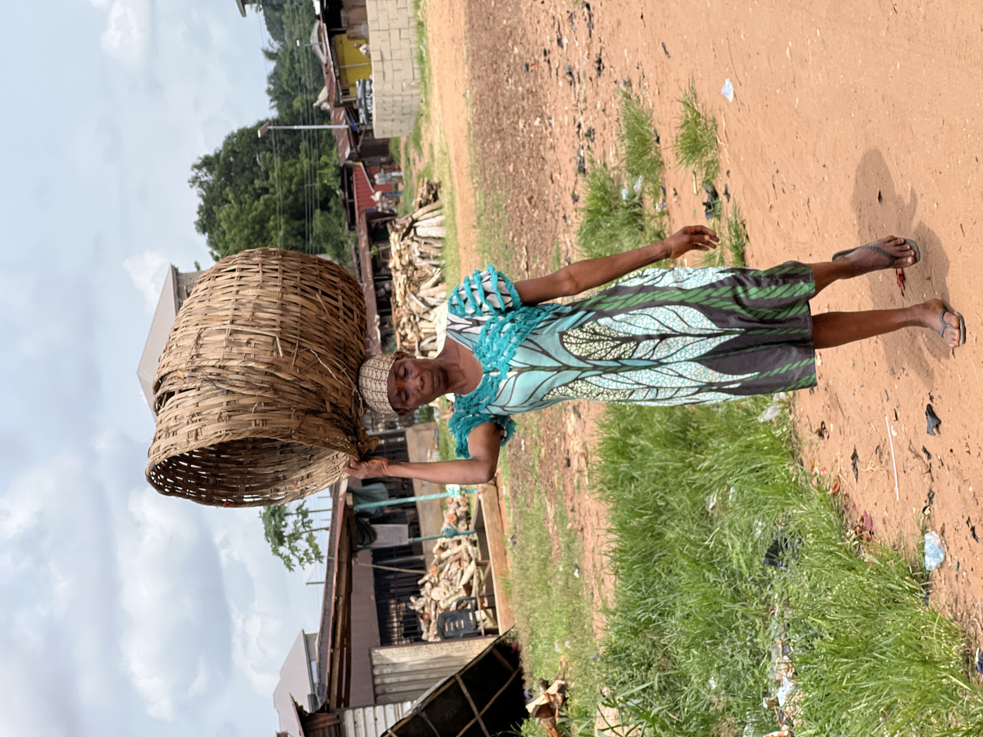 This full-body, portrait-oriented image, set in Obosi, Nigeria, captures an adult woman balancing a large, light-brown woven basket on her head as she stands on a dirt path. She is looking directly at the viewer with a neutral expression.
The woman is dressed in a vibrant, ankle-length dress that features a light blue base adorned with a striking pattern of green and black leaf-like motifs. Her short sleeves are light blue and appear ruffled. A light-colored headwrap or scarf covers her head. She wears simple dark-colored flip-flops on her feet. Her left hand is slightly raised, appearing to support the basket or provide stability, while her right arm hangs by her side.
The background consists of a wide, reddish-brown dirt road or path on the right, and areas of tall green grass interspersed with small amounts of trash on the left. Behind the woman, several rustic structures, possibly houses or sheds made of concrete blocks and corrugated metal, are visible. Piles of cut wood or logs are stacked near these buildings. Beyond the immediate structures, dense green trees and foliage extend into the distance, beneath a bright, overcast sky. Power lines and a street lamp are also discernable in the upper background. The natural, diffused lighting suggests it is daytime, likely under cloudy conditions. No discernible text is visible in the image.