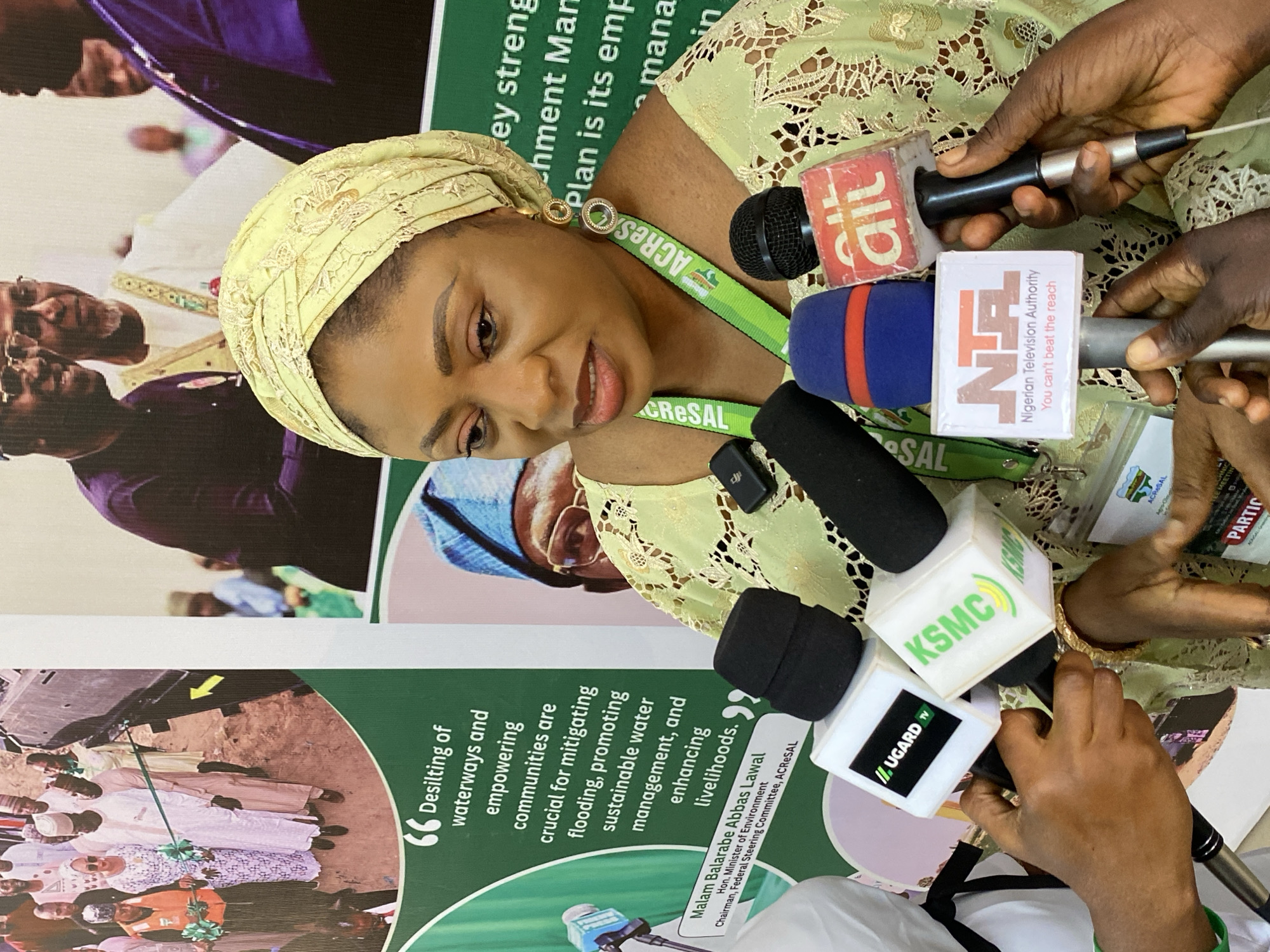 A woman in a light yellow-green lace top and a matching head covering is featured prominently, looking slightly downwards with a focused expression. She is surrounded by several hands holding microphones towards her, indicating a press conference or interview. She wears a green lanyard with "ACRESAL" printed on it.
The microphones display logos of various media outlets: AIT, NTA (Nigerian Television Authority, with the slogan "You can't beat the reach"), KSMC, and UGARD TV. The setting appears to be an indoor event, likely in Kwarau, Nigeria, given the visible Nigerian media outlets.
In the background, a large banner is partially visible, featuring text and images of other people. Notable text on the banner includes a quote attributed to "Malam Balarabe Abbas Lawal, Hon. Minister of Environment, Chairman, Federal Steering Committee, ACRESAL": "Desilting of waterways and empowering communities are crucial for mitigating flooding, promoting sustainable water management, and enhancing livelihoods." Other fragments like "ACRES" and "Agro-Olma" are also present on the banner, further indicating the context of the ACRESAL project. The image is taken from a slightly elevated perspective.