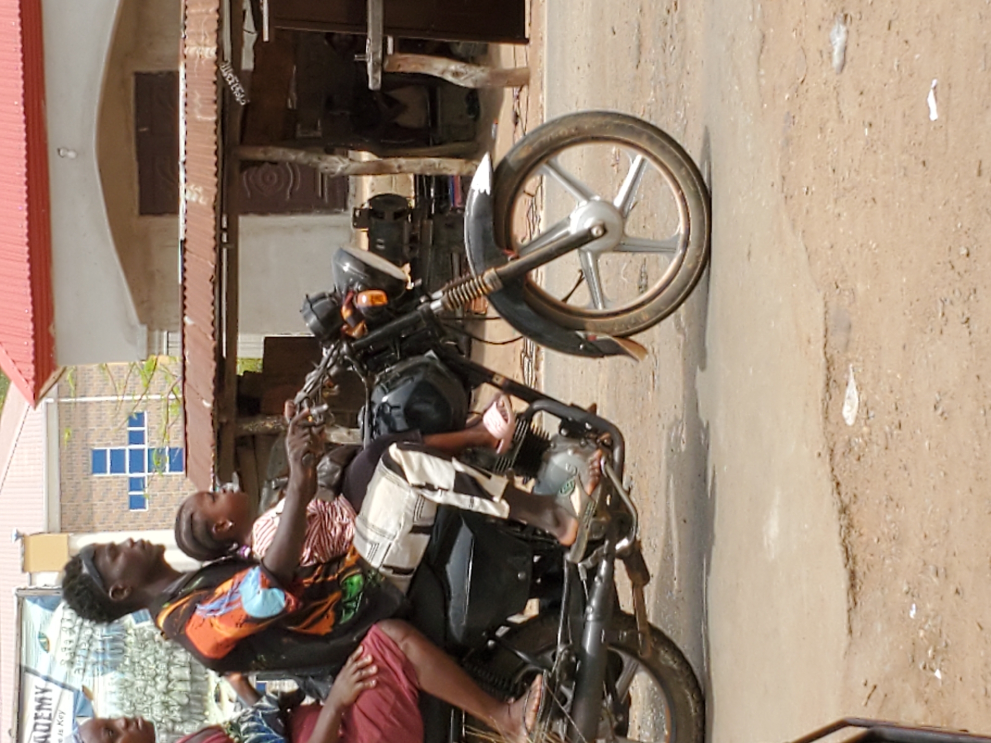 A medium shot captures a group of people on a motorcycle in Mararaba, Nigeria. Three individuals are visible: the rider, a young man with a black headband and colorful shirt, sits at the front. Behind him is a child in a red and white striped shirt and a skirt. A third person sits behind the child, wearing a maroon top. They are on a black motorcycle, parked on a dirt road. The front wheel of the motorcycle is prominent in the frame. Buildings are visible in the background. A building with a red roof and another with a blue cross design are also visible.