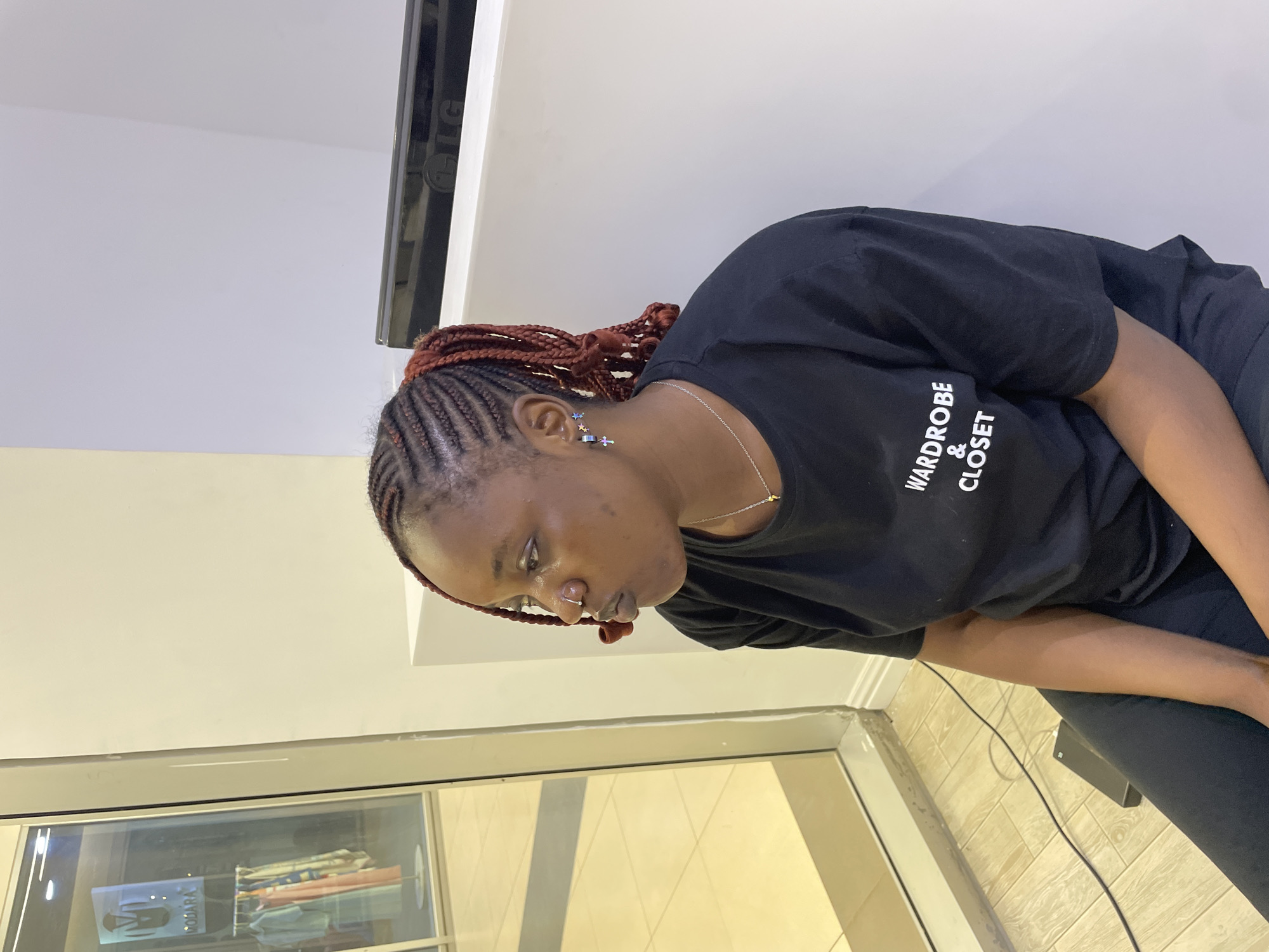 In Unguwar Sarki, Nigeria, a young Black woman with braided hair is seated indoors, looking to her left. She wears a black t-shirt with white text that reads "WARDROBE & CLOSET", a silver necklace with a small pendant, and earrings. One earring appears to be a star, and the other a cross with a star. She also has a nose ring. The woman is positioned in front of a white wall, and to her left is a large glass window reflecting an indoor store with clothing on display. The floor is tiled with a light-colored wood pattern. A black power bank is visible on the floor near her. The lighting suggests an indoor setting, possibly a retail space or a waiting area.