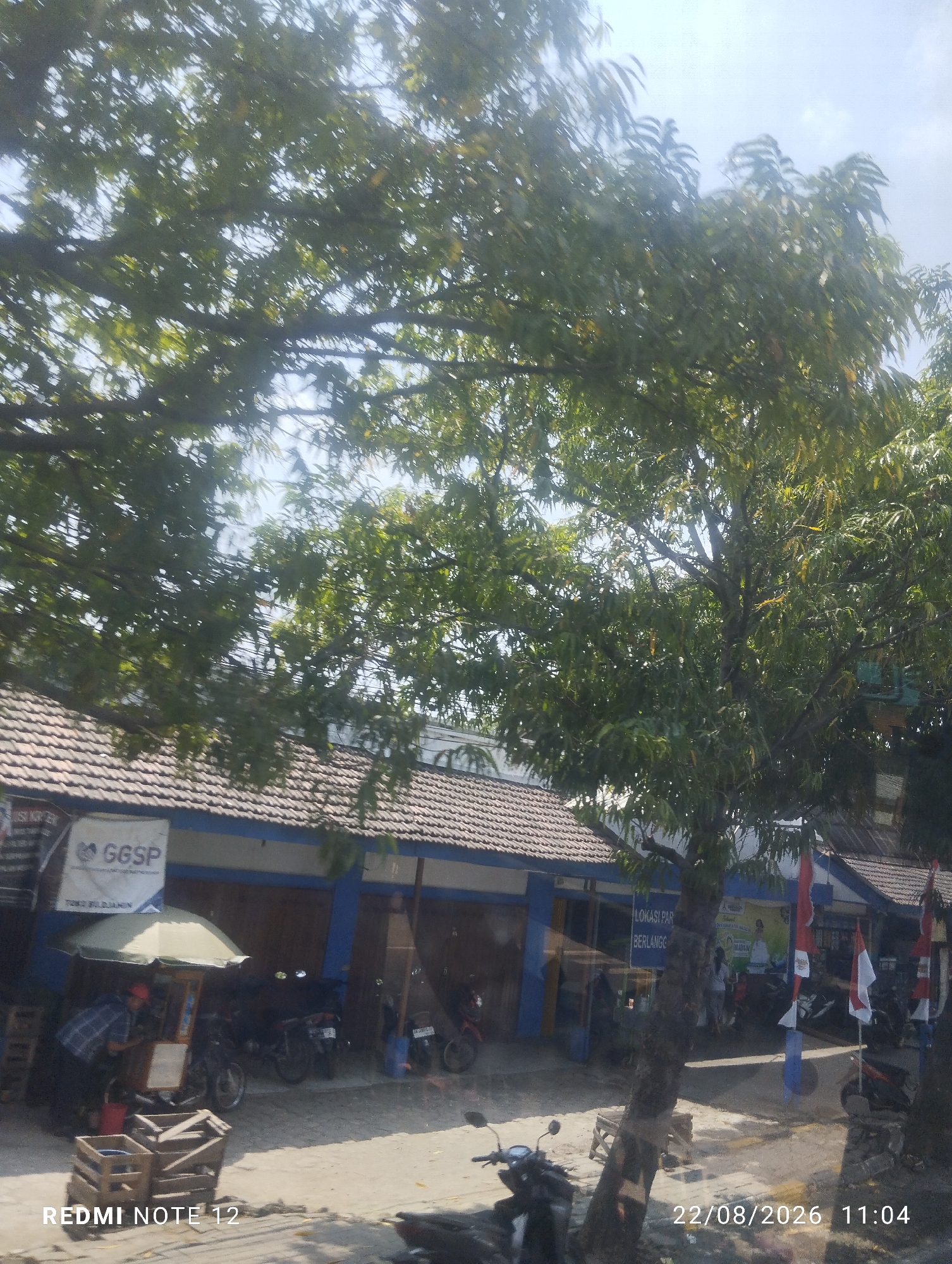 The image depicts a street scene in Kwangsen, Indonesia, on August 22, 2026, at 11:04 AM. The foreground is dominated by a large tree with lush green foliage, casting shadows on the street and buildings. To the left, a building with a tiled roof and blue trim is visible. A sign with the text "GGSP" is mounted on the building. In front of the building, a small food stall with a green umbrella is set up, and a person is interacting with it. Several motorcycles are parked in the vicinity. Further down the street, more buildings with similar architecture and tiled roofs are present. Flags are displayed on poles in front of one of the buildings. The street appears to be paved, and there are some wooden crates on the ground. The overall environment suggests a commercial or public area within the city.