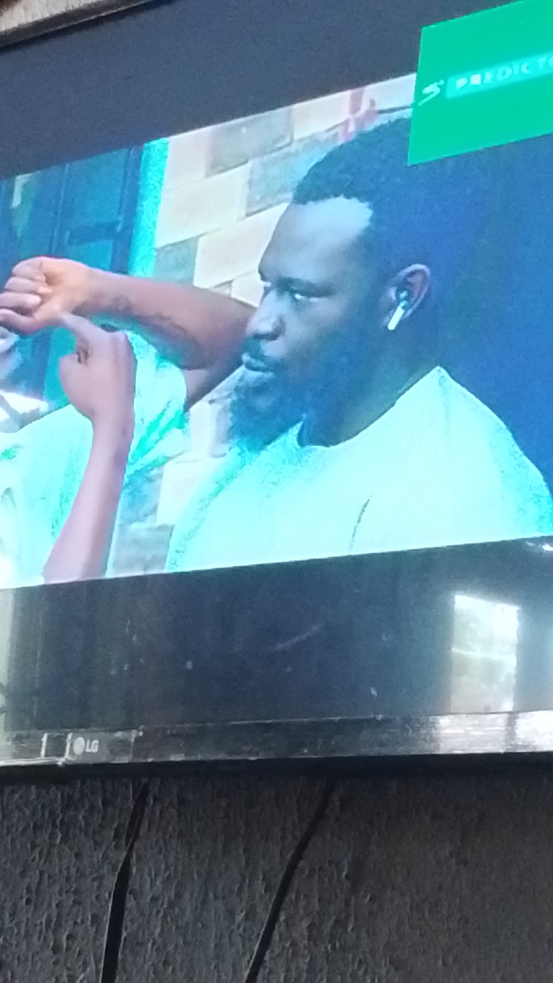 A close-up shot shows a man with short, dark, curly hair, a beard, and a light blue t-shirt wearing a white earbud. He is looking to the left. In front of him, another person's arm and hand are visible, with their index finger pointed towards the man. A green sign with white text is visible in the upper right corner of the image, partially obscuring the background. The background appears to be a wall with a brick pattern and a blueish-green light. The overall image is slightly blurry and captured from a low angle, suggesting it was taken on a television screen. The LG logo is visible on the bottom bezel of the screen, indicating the television's brand. This scene is likely taking place in Lokoja, Nigeria, given the context provided.