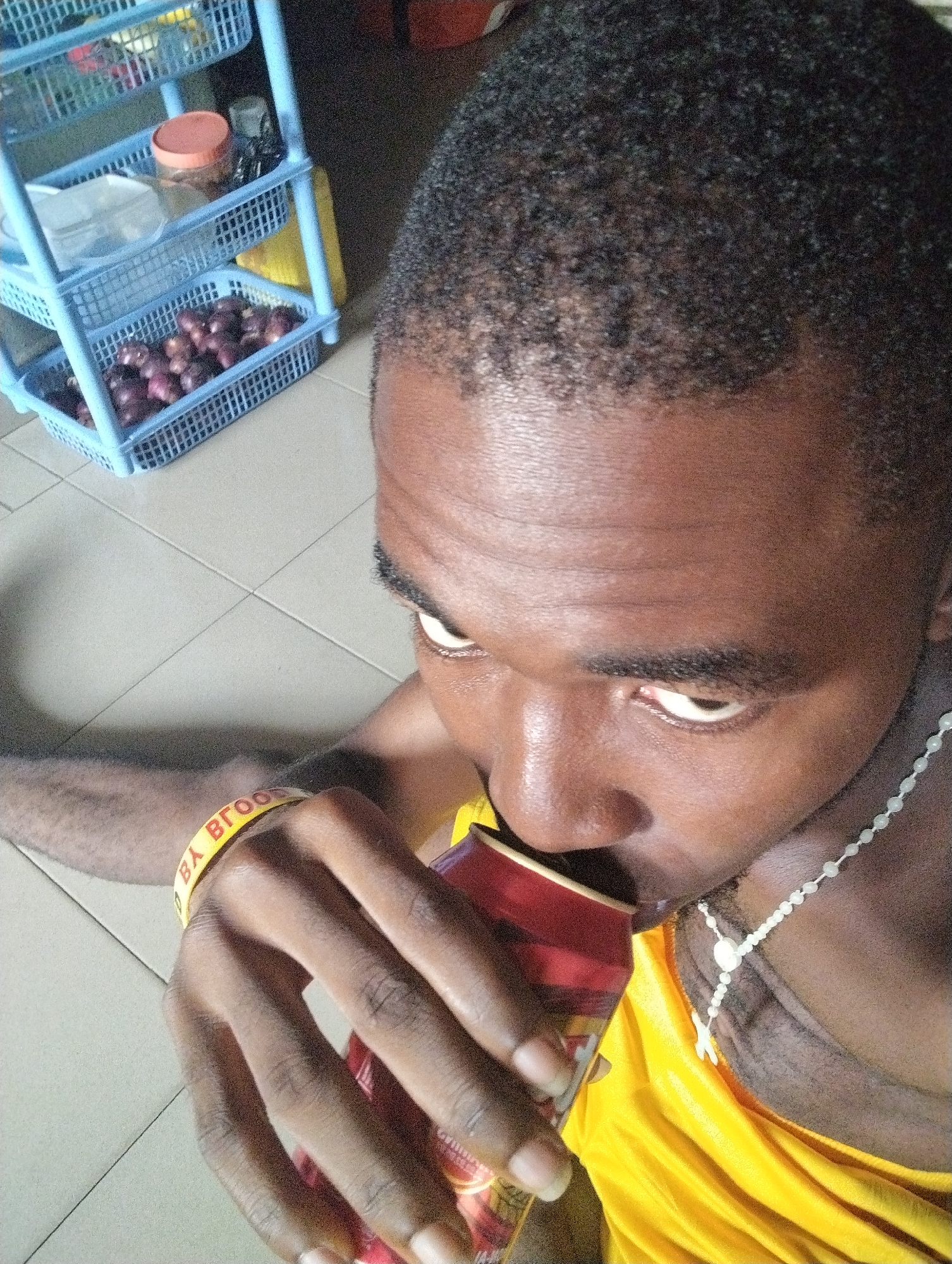 This close-up, high-angle photograph, likely taken in Ihiagwa, Nigeria, features a person (appearing to be male) drinking from a can in an indoor setting. The individual has dark skin and short, tightly-curled black hair. They are wearing a bright yellow t-shirt and a light-colored beaded necklace that includes a white cross pendant. A yellow wristband with red text, partially visible as "BY BROO", is on their right wrist.
The person is holding a red and gold beverage can and drinking from it, with their gaze directed upwards and slightly to the right. The floor beneath them is made of light-colored square tiles. In the background, a light blue, three-tiered plastic shelving unit is visible. The bottom shelf of this unit is filled with red onions. Other items, such as jars and plastic bags, can be seen on the higher shelves. The lighting suggests an indoor environment, and the overall scene depicts a casual moment within a household setting.