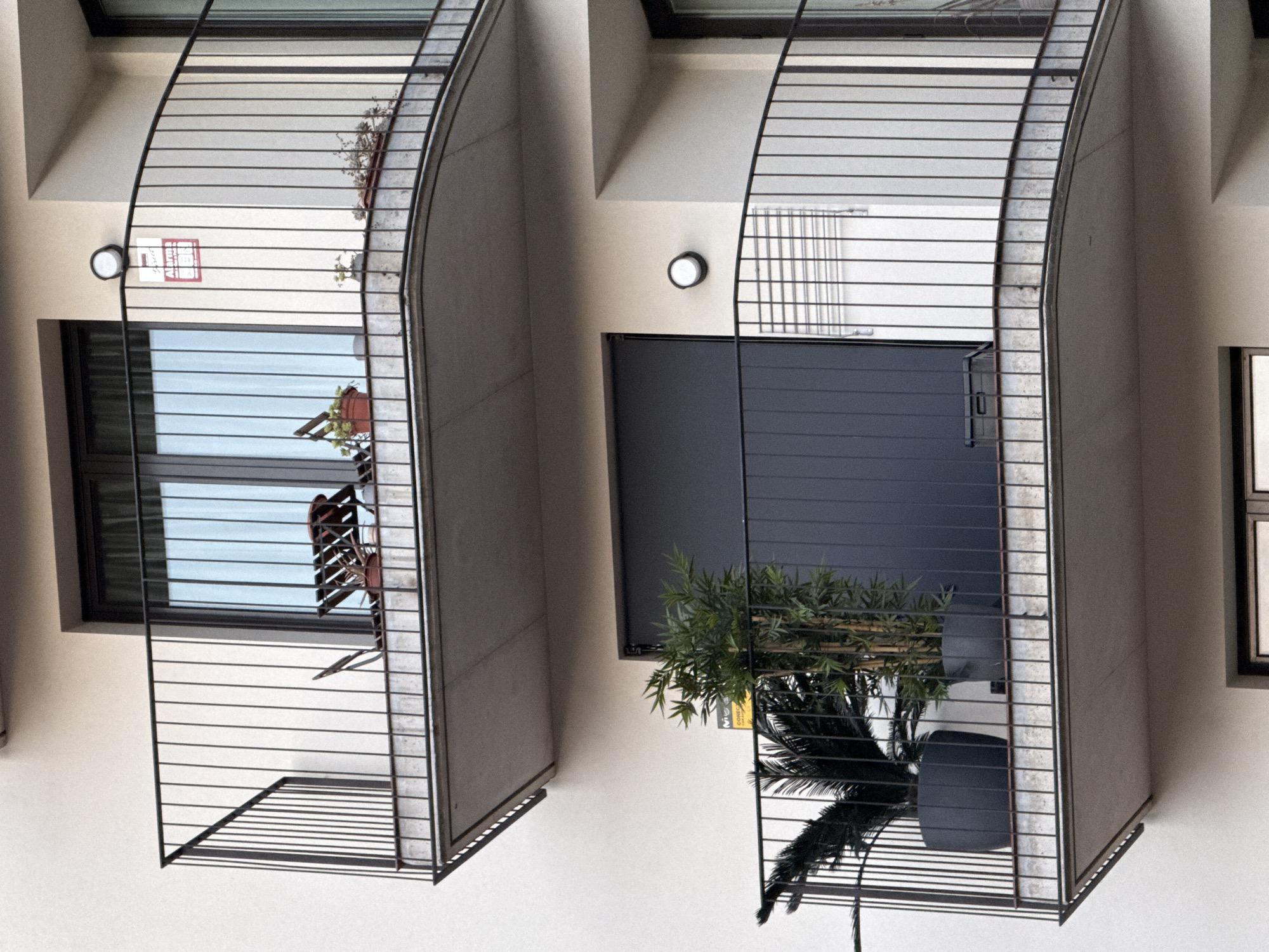 This media file shows an external view of two balconies of an apartment building, viewed from below. The balconies have curved metal railings and are attached to a light-colored building. The balcony on the left has a window with horizontal blinds and a potted plant on a stand outside it. Above the window, a round light fixture is mounted on the building wall, alongside a red and white rectangular sign with text.
The balcony on the right is wider and features a dark-colored door or window, with greenery and a palm frond visible behind the railing. A black plastic bin is placed on the floor of this balcony. A round light fixture is also visible on the wall of this section of the building. The architecture suggests a modern apartment complex. The lighting indicates it is daytime. The building appears to be in an urban setting, potentially a residential area in Barcelona, Spain, given the context provided. There is no discernible activity or emotional tone.
Visible text includes the word "Alarma" in red on the sign on the left balcony.