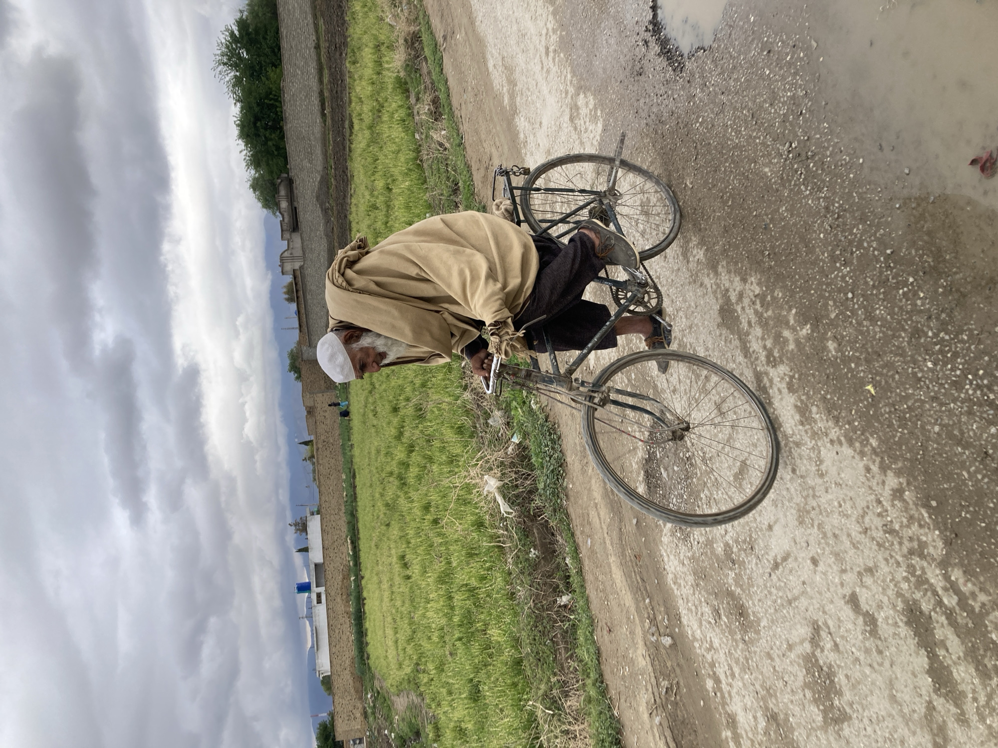 The image depicts an elderly man riding a bicycle on a dirt road in a rural area, likely in the Quetta City Tehsil, Pakistan. The man is wearing a white cap and a light brown shawl. The bicycle is old, with rusted components. The road is unpaved, with visible dirt and puddles. Adjacent to the road are fields of green crops and stone walls. The sky is overcast.