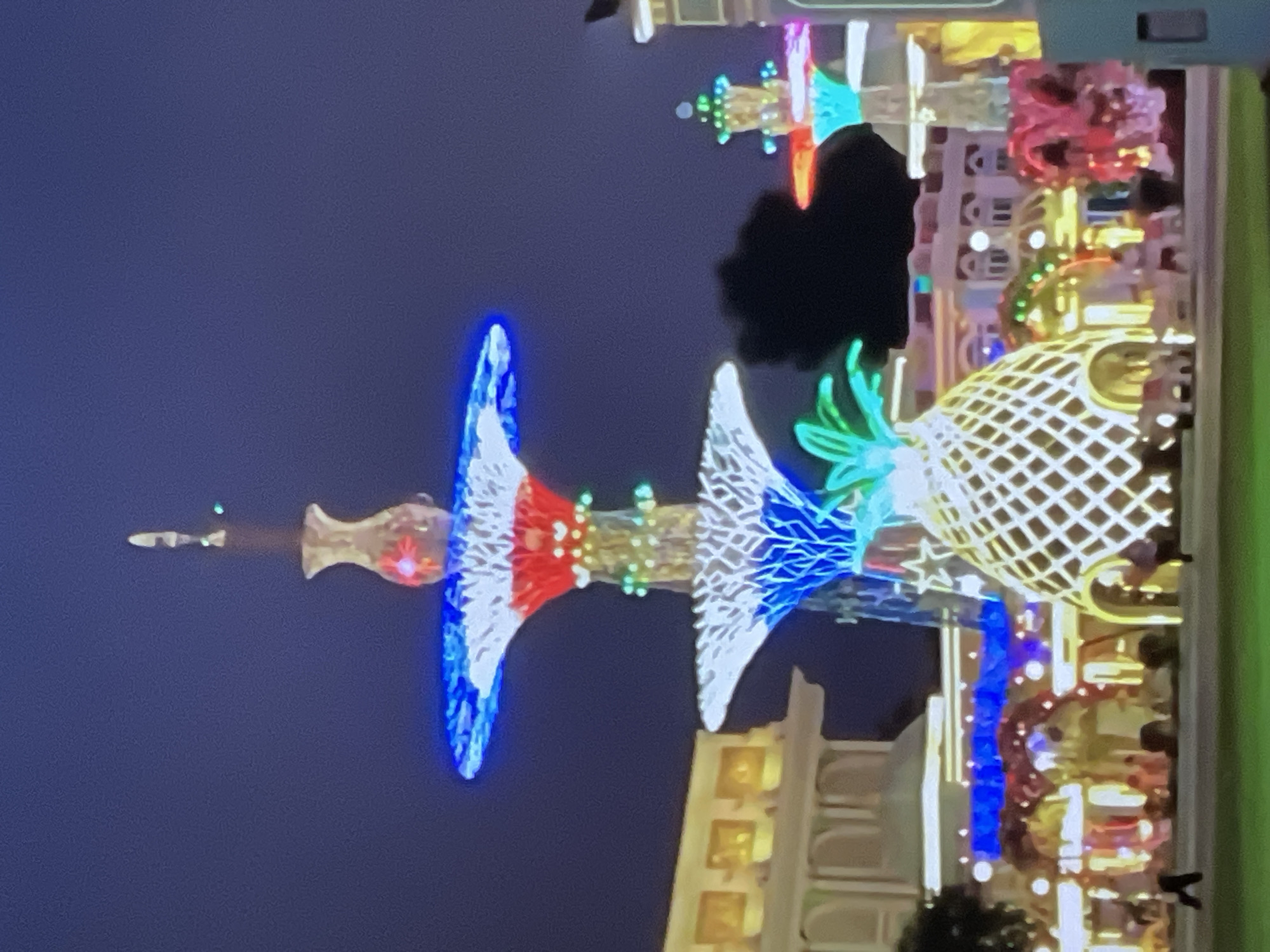 This image depicts a nighttime scene in Pattaya City, Thailand, featuring a brightly lit, multi-tiered decorative structure resembling a fountain or tree, adorned with white, red, and blue lights. The structure is topped with a vase-like element. At the base, a large illuminated pineapple-shaped decoration with green leafy fronds is prominent. Behind these central features, several buildings with illuminated windows and arched entrances are visible, suggesting a commercial or entertainment district. The sky is dark blue, indicating evening or nighttime. Several figures, likely people, are dimly visible in the foreground and middle ground, walking or standing. The overall scene suggests a festive or decorative display.