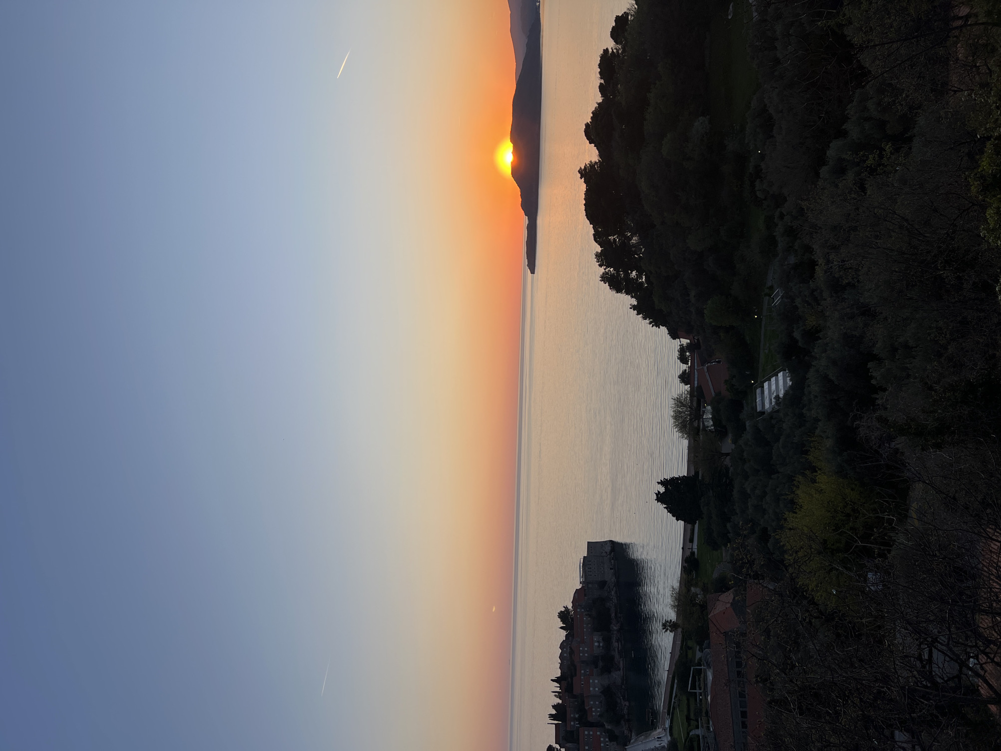 The image depicts a sunset scene in Sveti Stefan, Montenegro. The sun is setting behind a mountain range across the water, casting an orange glow across the sky and sea. The island of Sveti Stefan is visible in the foreground, with its distinctive red-tiled roofs and stone buildings. Lush vegetation covers the coastline on the right side of the frame. Two contrails are visible in the upper left of the frame.