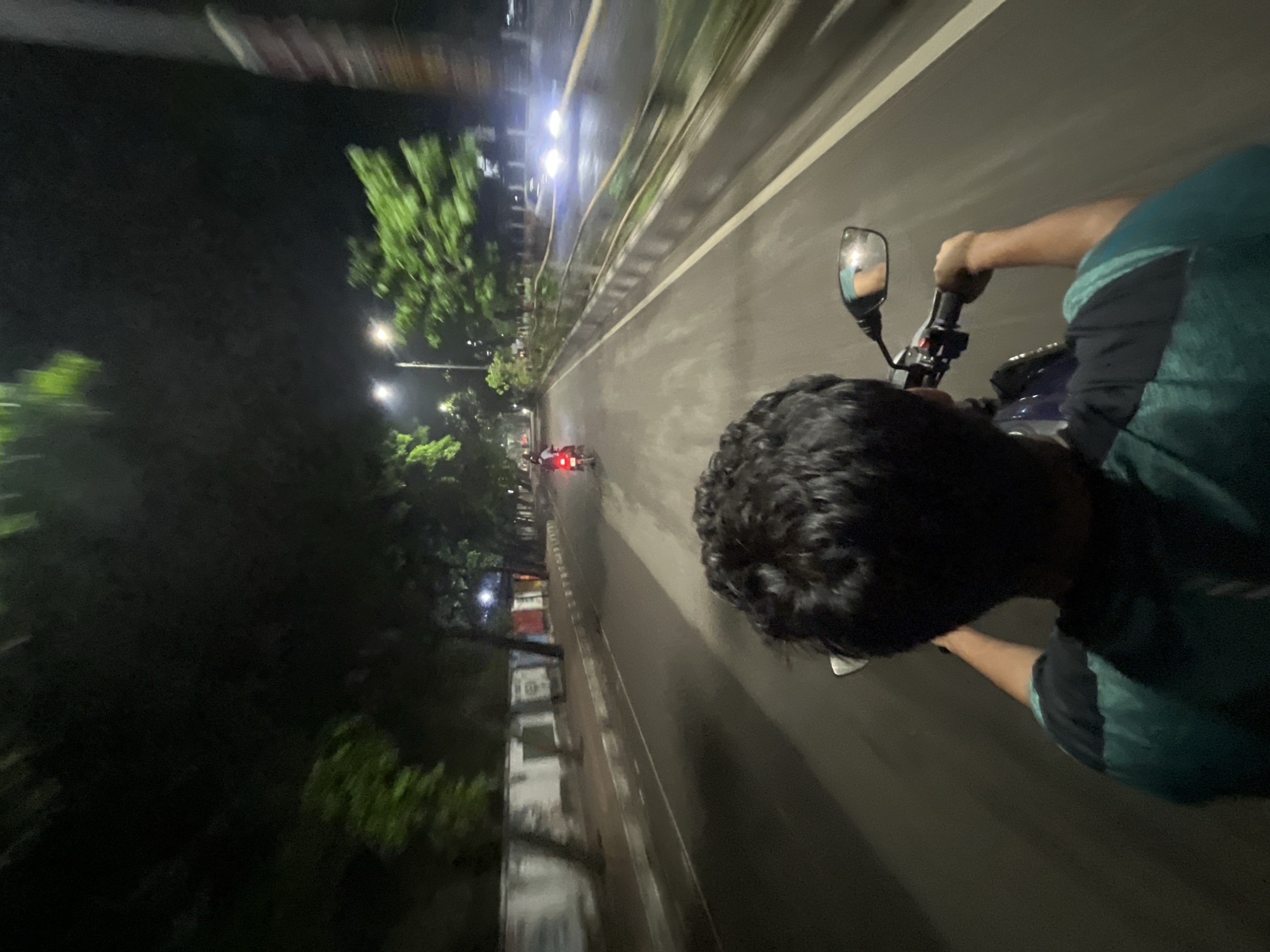 A person is riding a motorcycle along a road at night, likely in Dhaka, Bangladesh. The road is wet and reflects the streetlights. Trees line the side of the road. There is another motorcycle with red taillights ahead in the distance. The rider is wearing a dark-colored shirt with a green accent. The motorcycle's side mirror is visible.