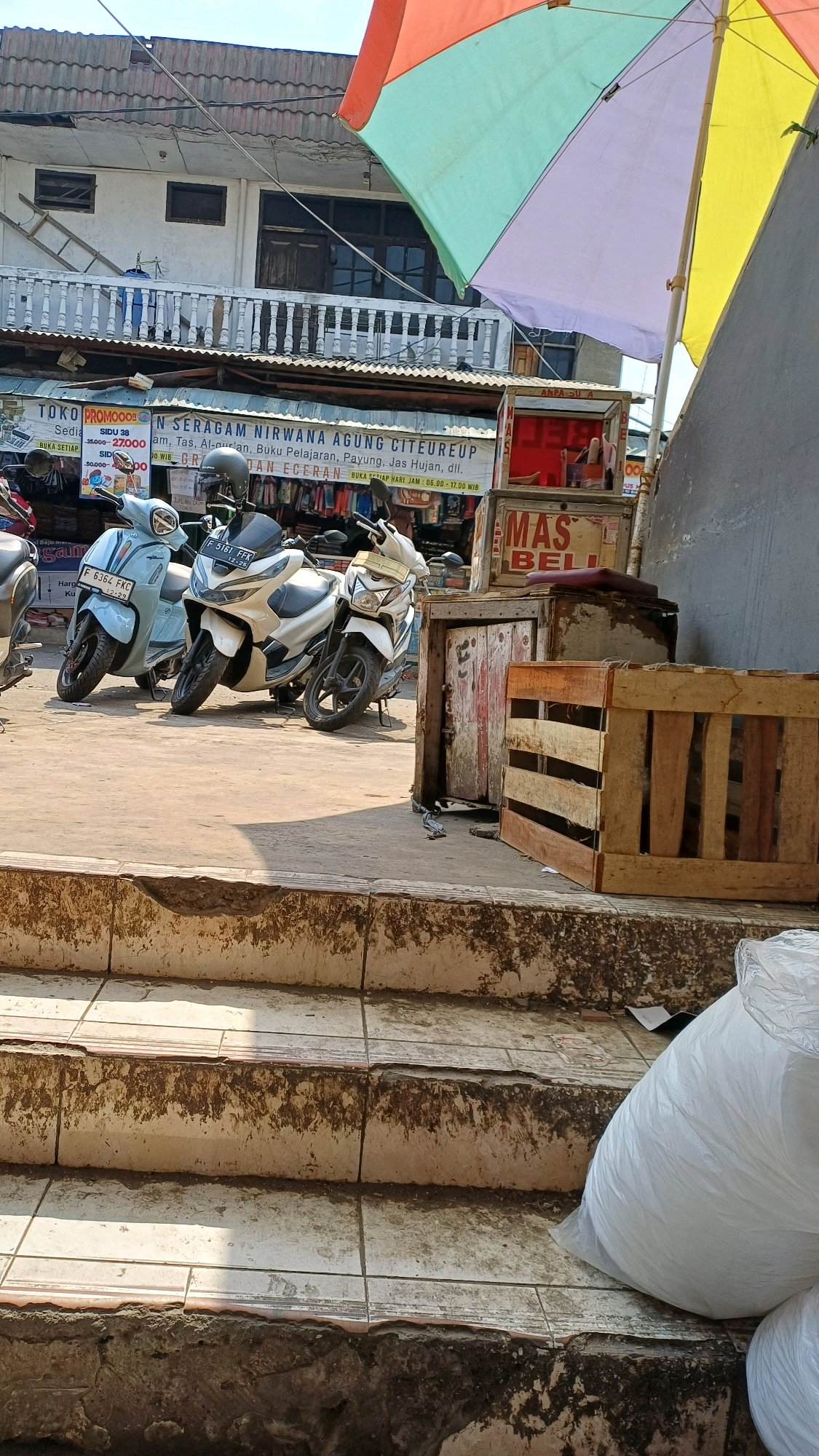 The image depicts an outdoor scene in Citeureup, Indonesia, likely a street or market area. In the foreground, a set of worn, tiled stairs leads upwards, with the lower steps showing signs of dirt and damage. To the right of the stairs, a large white plastic bag, possibly containing goods or trash, is partially visible.
Further back, a paved area is occupied by several motorcycles parked side-by-side. Three distinct motorcycles are visible: a light blue scooter on the left, a white Honda PCX in the center, and a white motorcycle with a black seat on the right. Behind the motorcycles, a row of stalls or shops can be seen. A prominent sign for "TOKO PROMOOO" is visible, followed by "N SERAGAM NIRWANA AGUNG CITEUREUP," indicating a clothing and uniform store. Text below suggests they sell school uniforms, student uniforms, school bags, umbrellas, raincoats, etc. Another sign reads "MAS BELI". Wooden crates are stacked near one of the stalls.
Above the stalls, a building with a tiled roof and a balcony with railings is visible. A colorful, large umbrella, divided into sections of red, orange, yellow, green, and purple, is open and extends over part of the scene, providing shade. The environment appears to be a typical bustling urban setting with elements of commerce and daily life. The lighting suggests it is daytime, with shadows cast by the umbrella and the building.