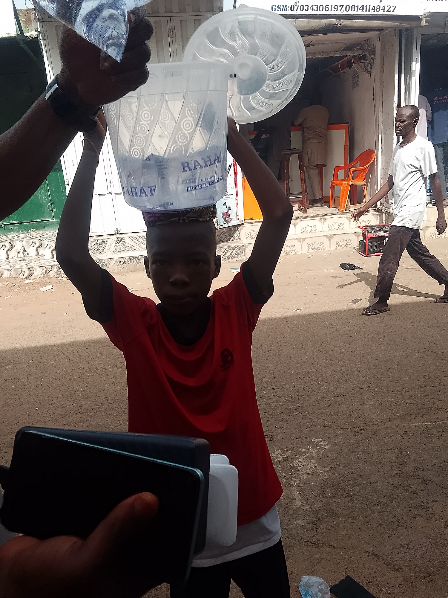 The image shows a young boy in the foreground wearing a red t-shirt, balancing a plastic water container on his head. Another person is pouring a sachet of water into the container. A mobile phone is visible in the lower left corner, held by someone taking the picture.
In the background, a man walks towards the right. Behind him is a building with text "GSM: 07034306197,08141148427" visible above the entrance. An orange chair and other figures are indistinctly visible inside the building. A generator sits on the ground near the building.
The brand name on the water container is "RAHA" with the text "HAFDAC REG/NO/B1/714."
The location is suggested to be in the city of Gombe, Nigeria. The ground appears to be sandy or dusty.