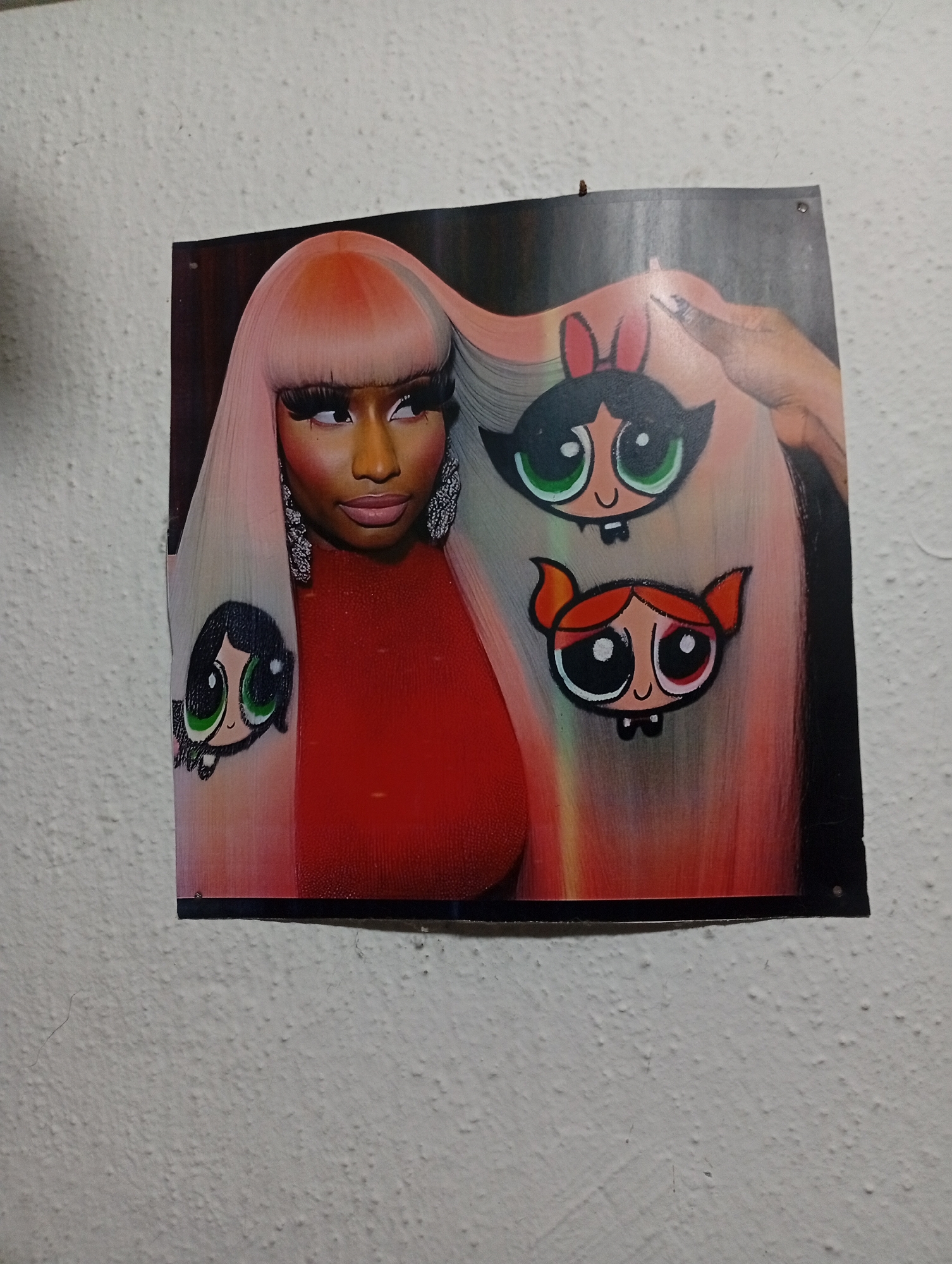 A square poster hangs on a textured, light-colored wall. The poster features Nicki Minaj in a red top with long, light pink and orange hair. The Powerpuff Girls characters Buttercup, Blossom, and Bubbles are drawn on her hair. A hand with manicured nails is touching the hair. No indications of a specific location in Lokoja, Nigeria, are visible in the image.