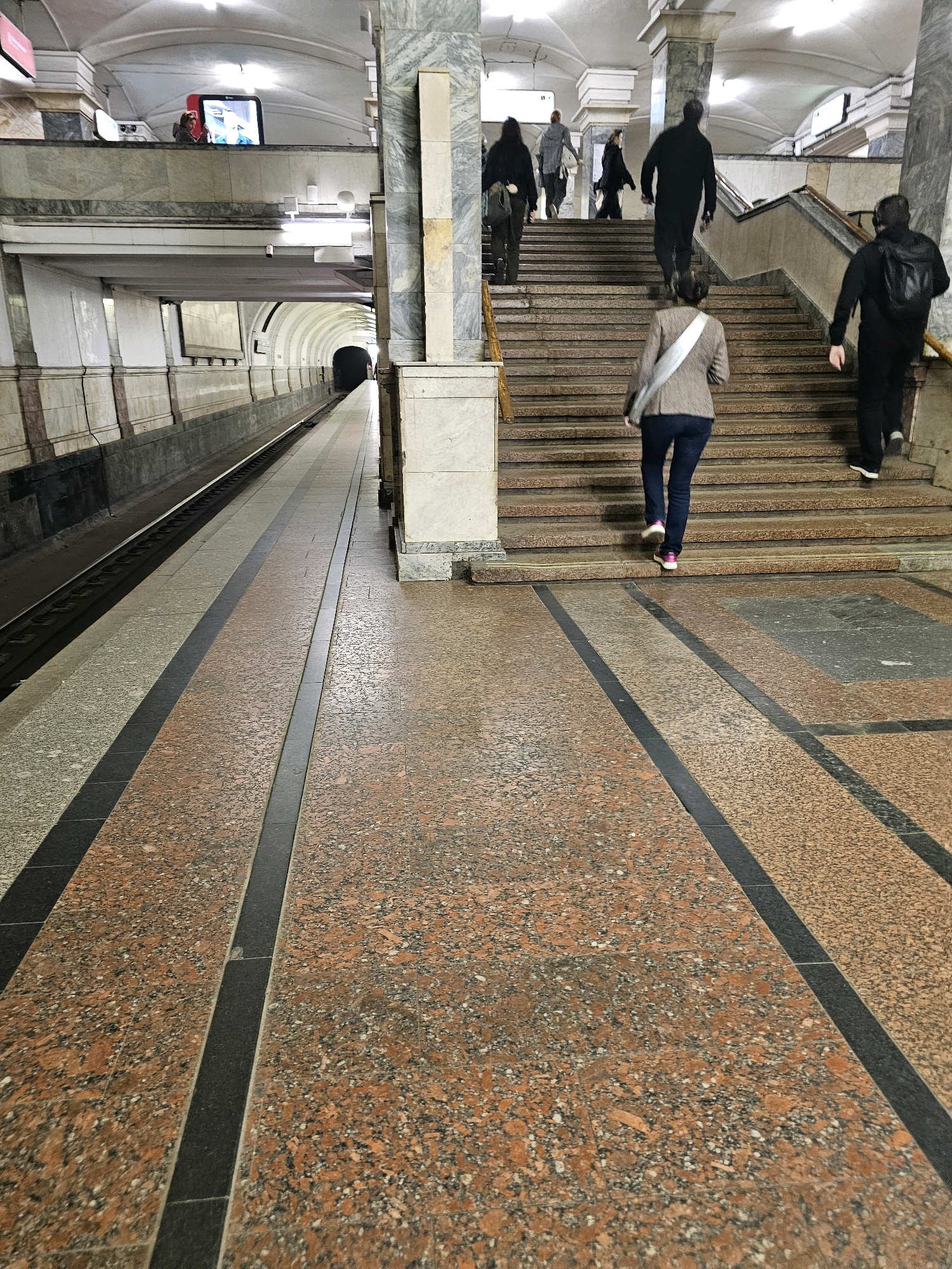 This is a photograph of a subway station in Moscow, Russia. In the foreground, a woman in a brown jacket and dark jeans is walking up a wide staircase, her back to the camera. She has a light-colored sash draped over her shoulder. To her right, a person wearing a black jacket and backpack is also ascending the stairs. Further up the stairs, several other individuals, mostly obscured from view, are also walking upwards. The stairs are made of stone and are well-lit by overhead lights.
To the left of the stairs, a subway platform extends into the distance, with train tracks running alongside it. The platform is a mixture of light gray and reddish-brown speckled granite flooring, bordered by black strips. A tunnel entrance is visible in the background on the left. The architecture of the station is grand, with marble columns and high arched ceilings. On an elevated walkway to the far left, a person is visible near a screen displaying an image. The overall scene depicts a busy, functional subway environment.
