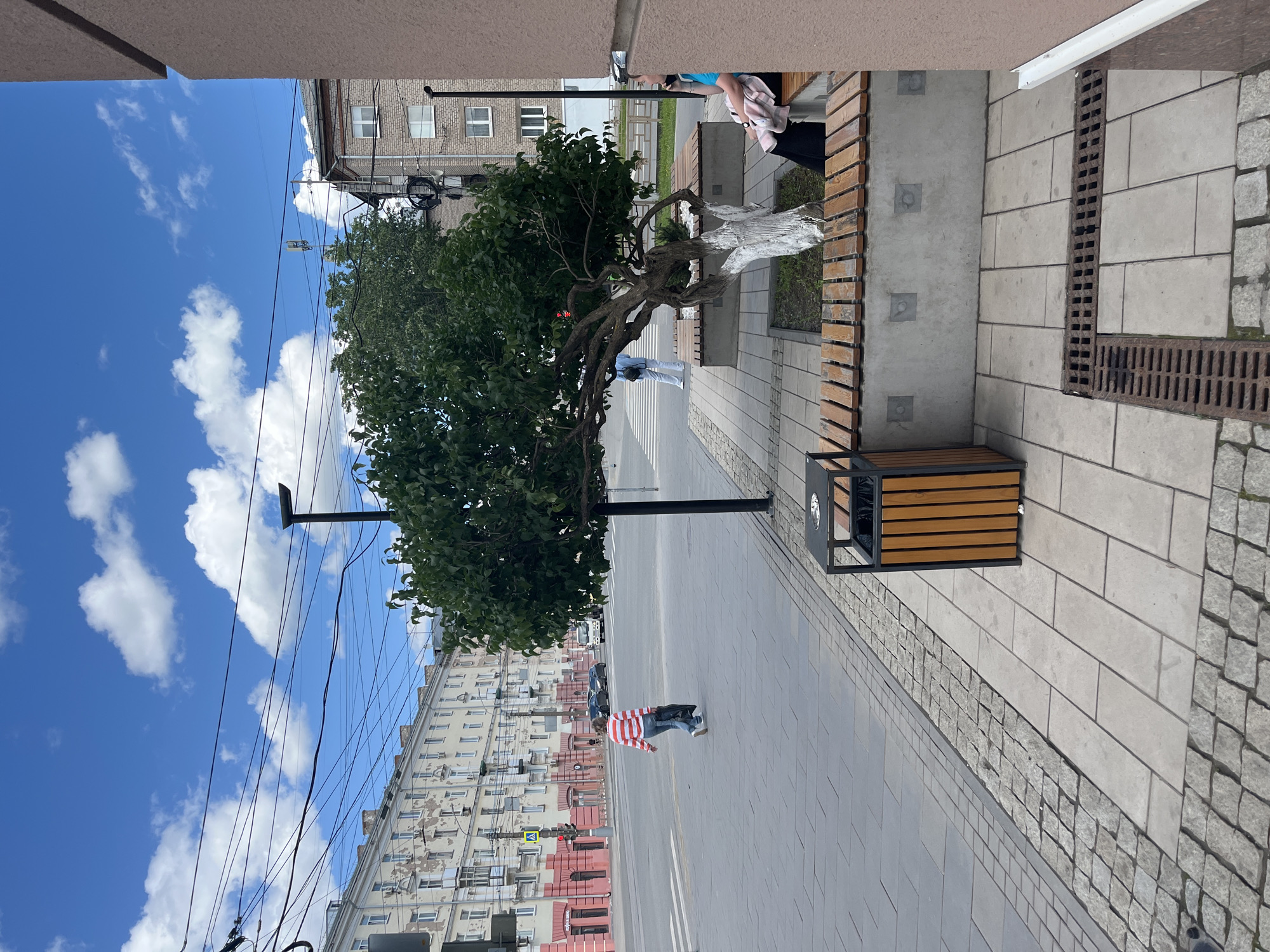 The image depicts a street scene in Izhevsk, Russia, showcasing an urban landscape. The street is paved with gray concrete slabs and a strip of granite cobblestones. A man in a red and white striped shirt and blue jeans is walking on the sidewalk.
A distinctive tree with a whitewashed trunk stands near a modern wooden bench and a metal-framed trash bin. The tree's branches are full of green leaves. Overhead, power lines crisscross the blue sky with scattered white clouds.
In the background, multi-story apartment buildings are visible. These buildings are painted in a pale color with reddish-brown architectural details. The sky is bright blue with scattered white clouds.