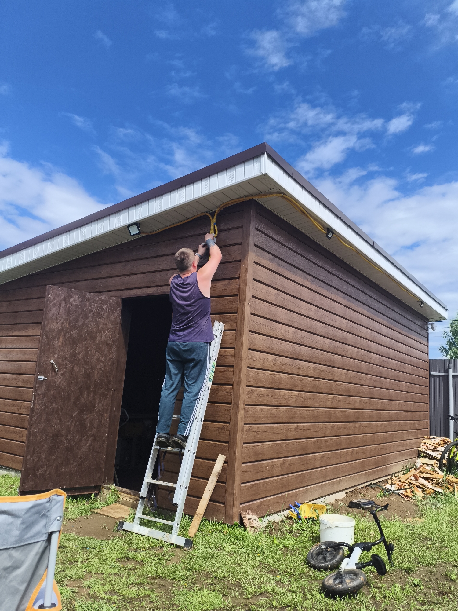 A man stands on a ladder attached to a brown shed, appearing to work on an electrical wire running along the building's upper edge. The shed has a white and brown roof, and an open door revealing its interior. A tricycle, a bucket, and various tools are scattered on the grassy ground surrounding the shed. A partial view of a folding chair, firewood, and a fence are also visible. The sky above is blue with scattered clouds. Based on the surroundings and the name of the location, the photo was most likely taken in a residential area near Русский Вожой, Russia.