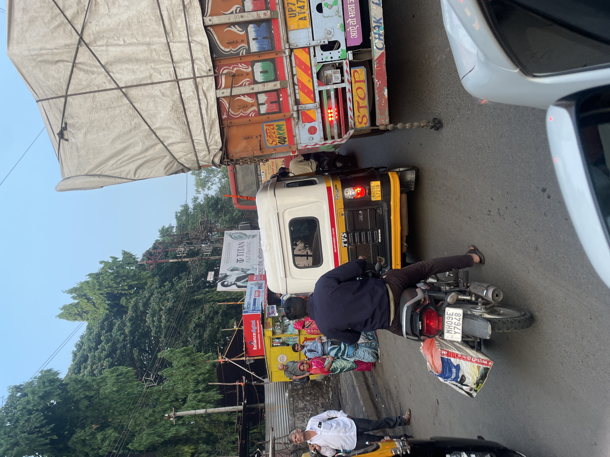 The image depicts a busy street scene in Kolhapur, India, identifiable from the "दसरा चौक, कोल्हापर" text on a Titan Automatic advertisement. A large cargo truck, with "STOP" prominently displayed on its rear, is chained to a white and yellow auto-rickshaw. A motorcyclist in a dark blue jacket is positioned between the auto-rickshaw and the right side of the frame. A man in a white shirt and dark pants stands near another yellow auto-rickshaw. A group of people, including some women in sarees, are near a Reliance Digital advertisement. Green trees and power lines are visible in the background, beneath a clear sky. The motorcycle has the license plate "MH09E Y7648". A bag is attached to the rear of the motorcycle. The auto-rickshaw has the license plate "MH05E L7218".